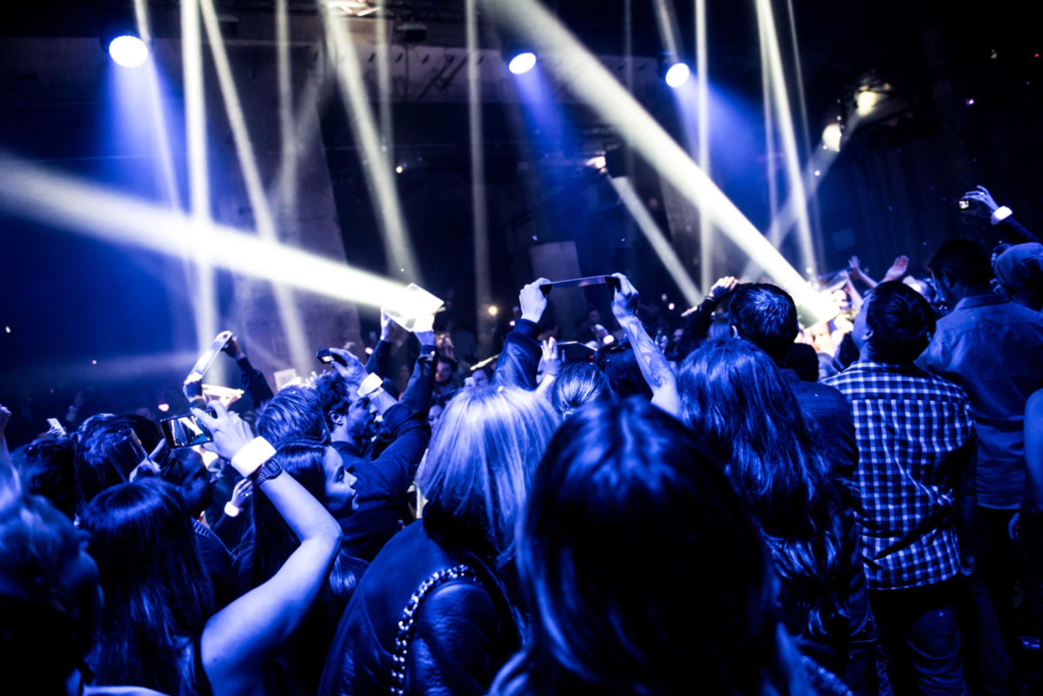 Performers in a crowd in a club environment reflect beams of spotlights from handheld mirrors.