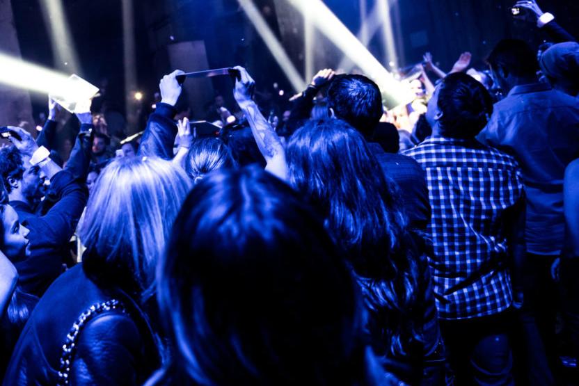 Performers in a crowd in a club environment reflect beams of spotlights from handheld mirrors.