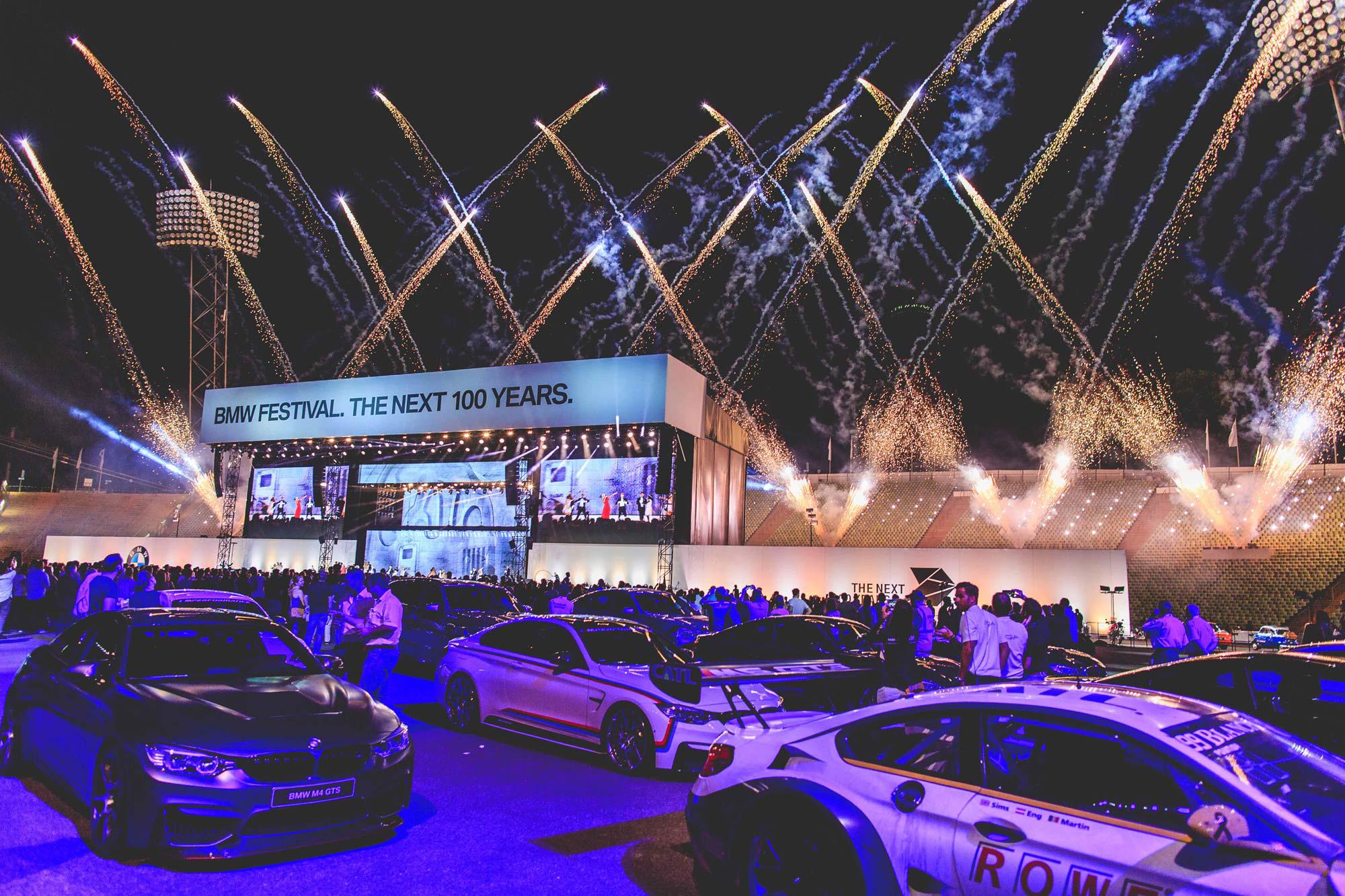 A fleet of BMWs and a crowd behind them. Fireworks burst into the sky and a large stage has a sign that says "BMW FESTIVAL. THE NEXT 100 YEARS." on it.