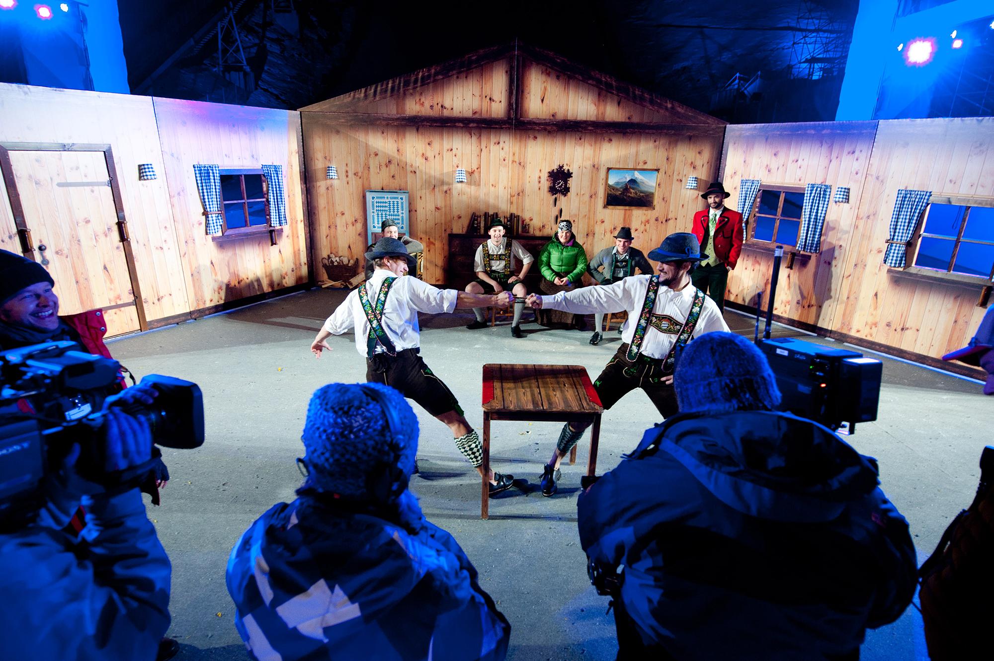 Performers on a stage set up to look like an alpine hut, wearing traditonal Tracht clothing, dance while a cameraperson films them.