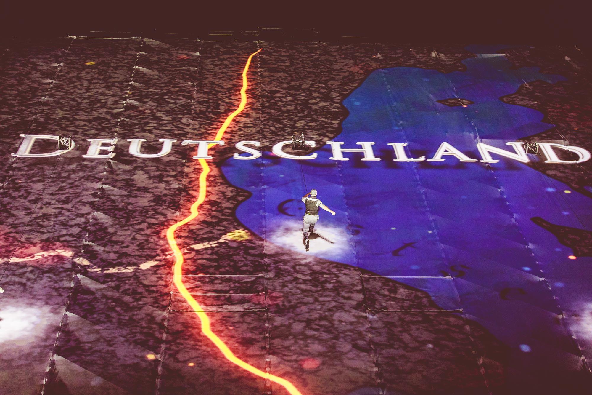 A performer runs up a large vertical wall with "DEUTSCHLAND" video mapped onto it.