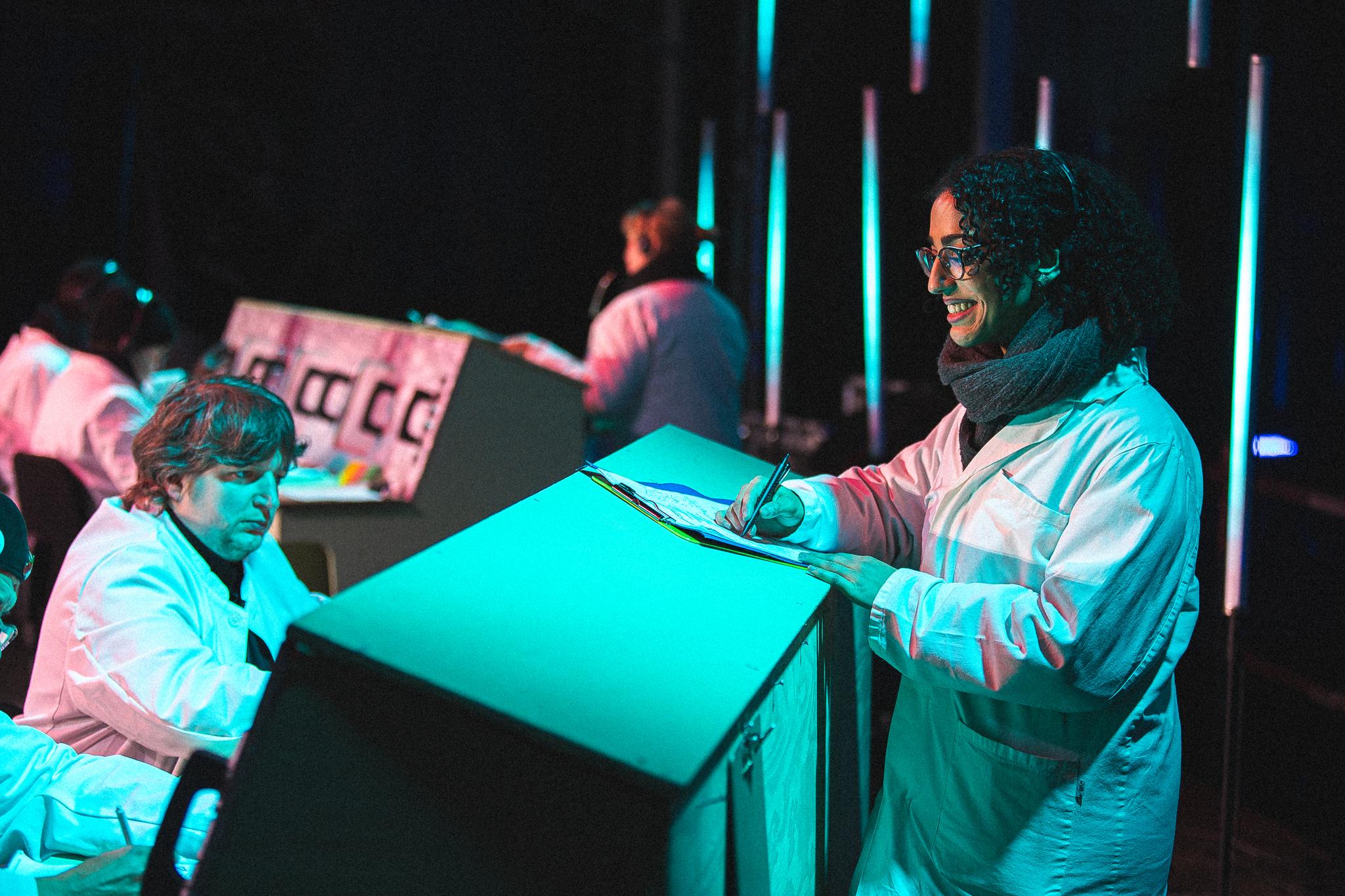A woman in a lab coat smiles as she writes something on a clipboard. More people in lab coats operate a chunky looking grey machine.