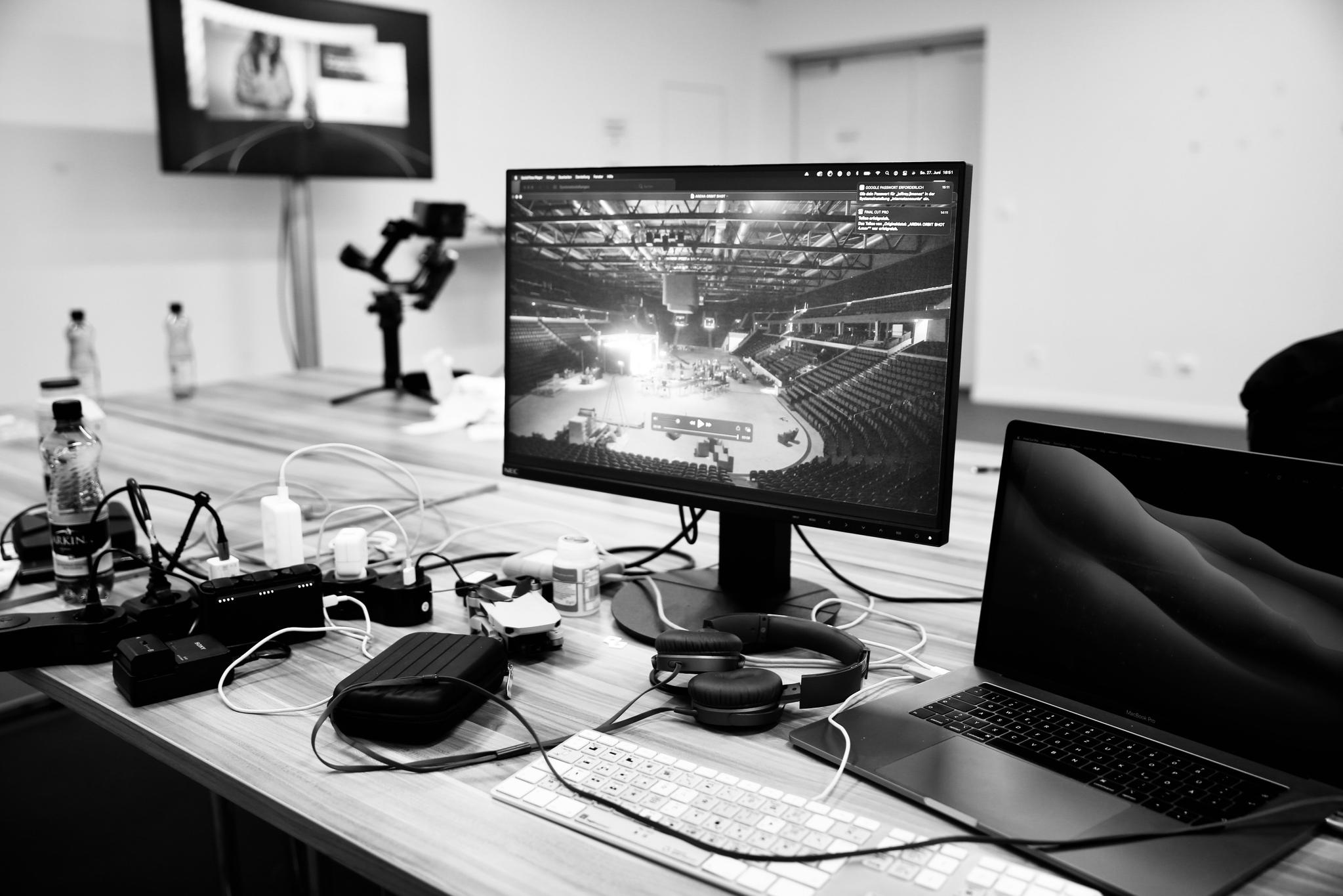 A black and white photo of 3 monitors: tv, computer, and laptop on a table in a nondescript white walled room. On the computer monitor there is footage of an empty stadium.