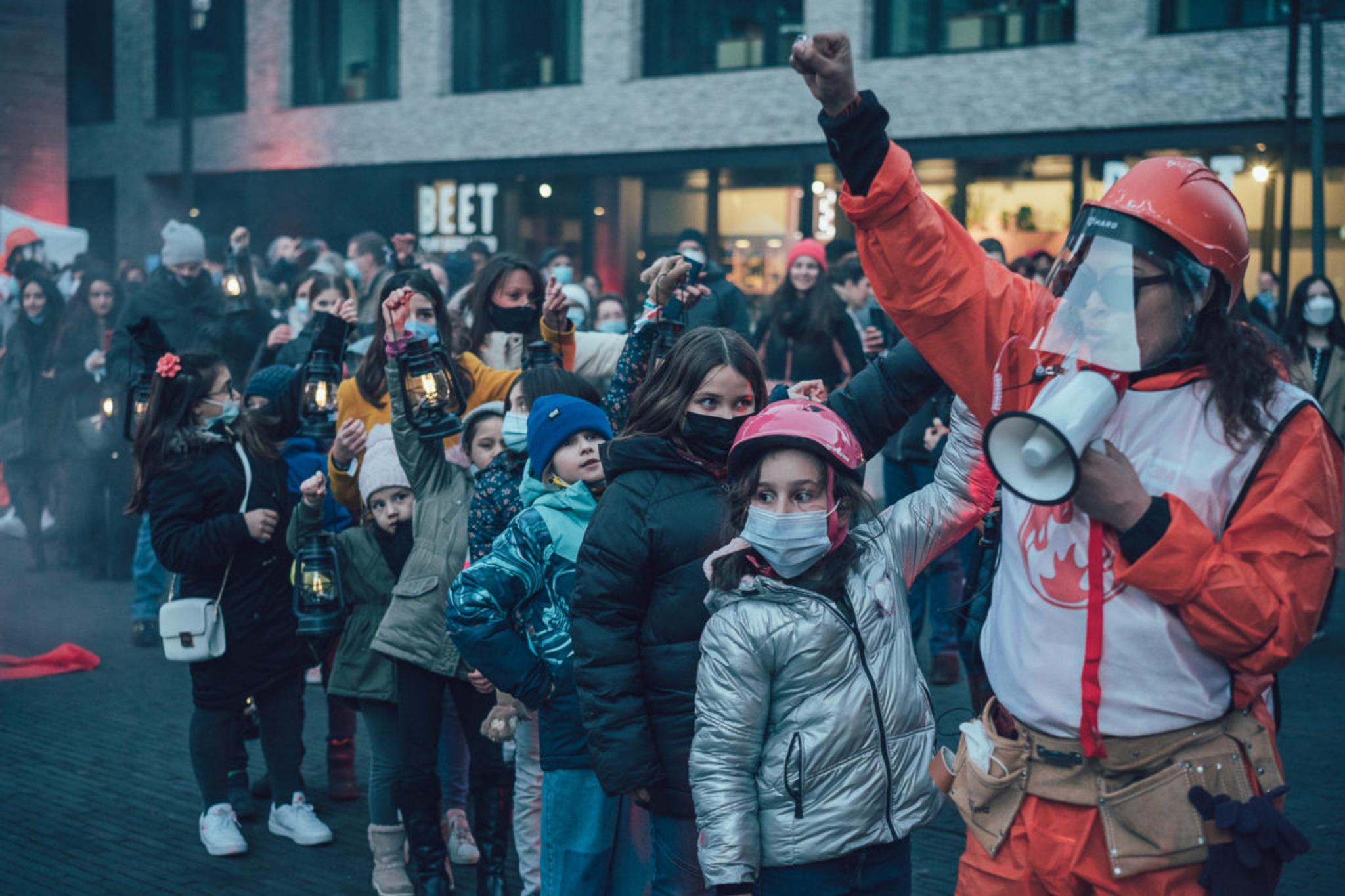 A woman in an orange jumpsuit and a white vest with a fire symbol on it has her fist in the air and is speaking into a megaphone. Children line up behind her, some wearing masks. They are outside a brown brick building in what looks like a plaza.
