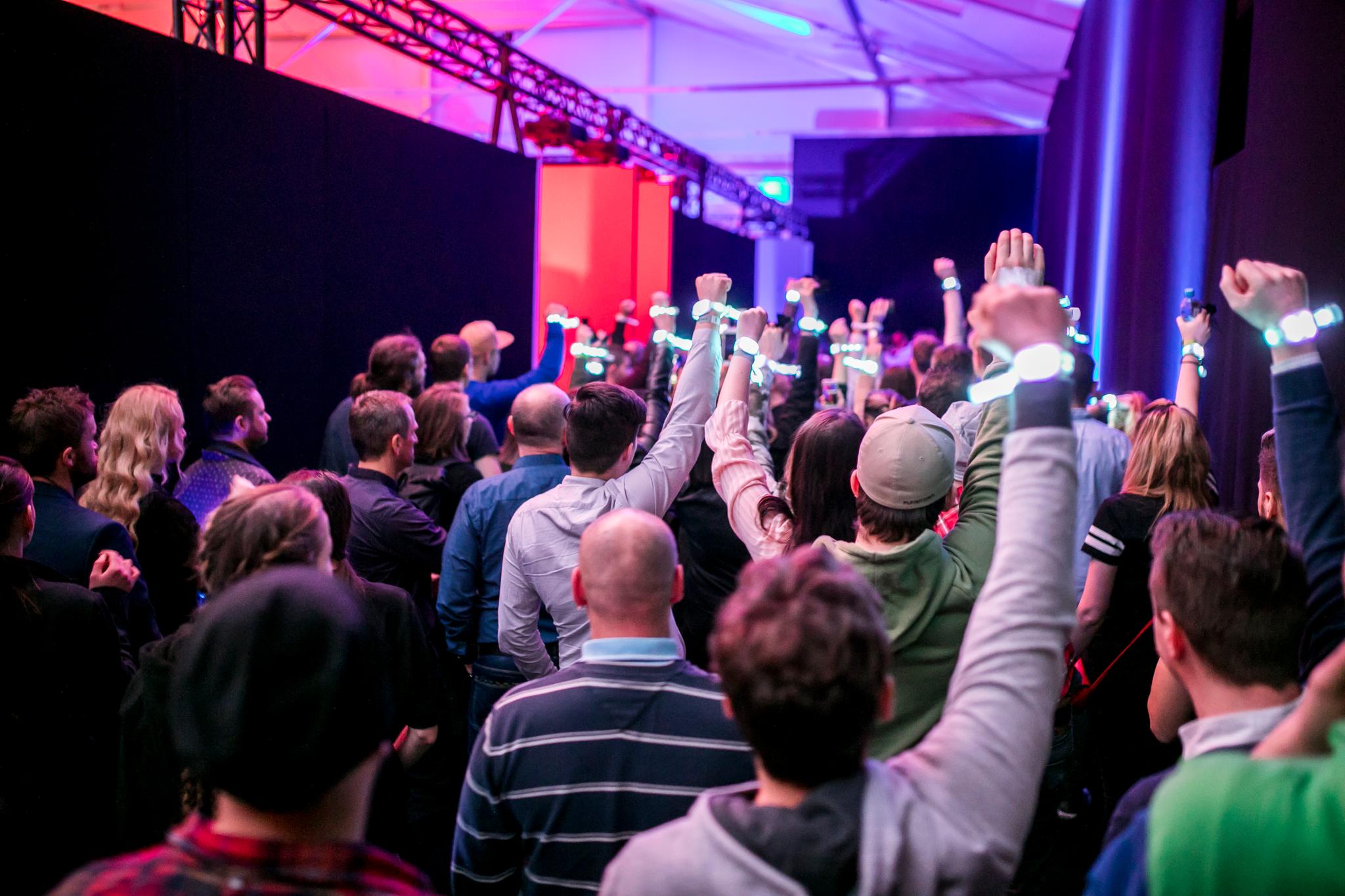 A crowd of people with their backs turned to the camera hoist their fists into the air- they are all wearing glowing white-blue wristbands. Colorful event lighting and trusses can be seen in the background.