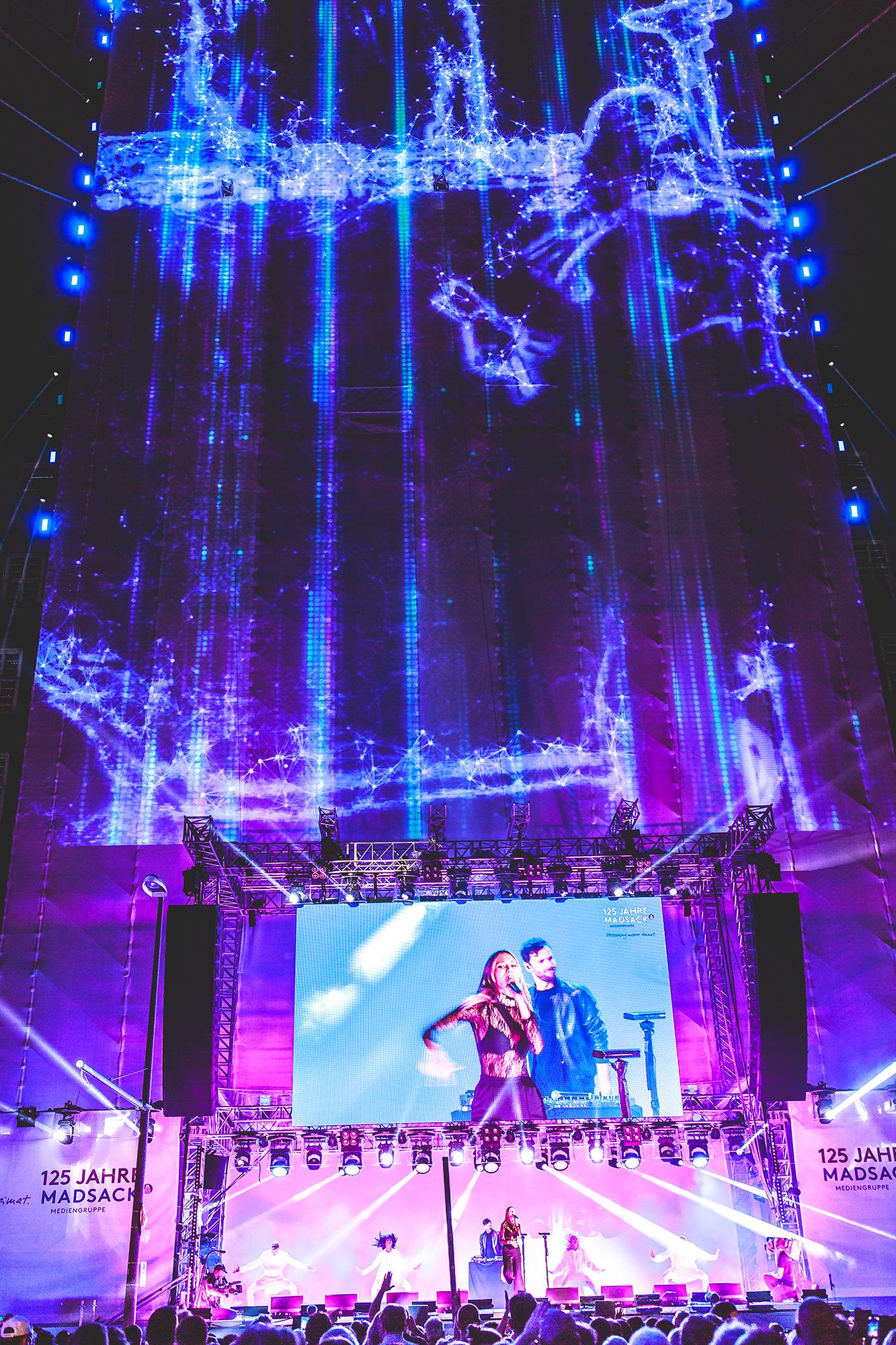 Video mapping of bright, blue sprite-like shapes on a large vertical stage. A large crowd watches a musician outdoors.