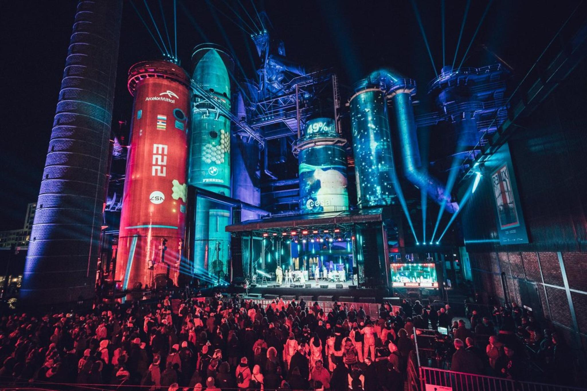 A wide shot of a crowd facing an outdoor stage. Blue lights are beaming out into the night sky, and industrial brick tube-towers and machinery frames the stage and has video content projected onto them.