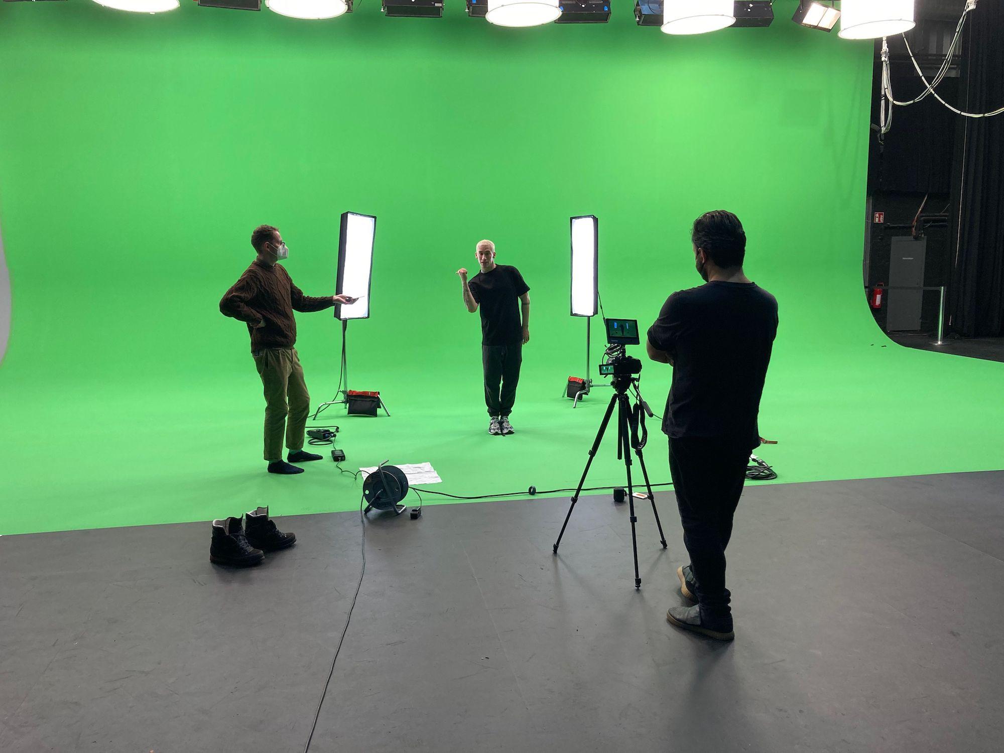 3 men stand in a green screen studio. One directs, one films, and one dances between 2 soft boxes.