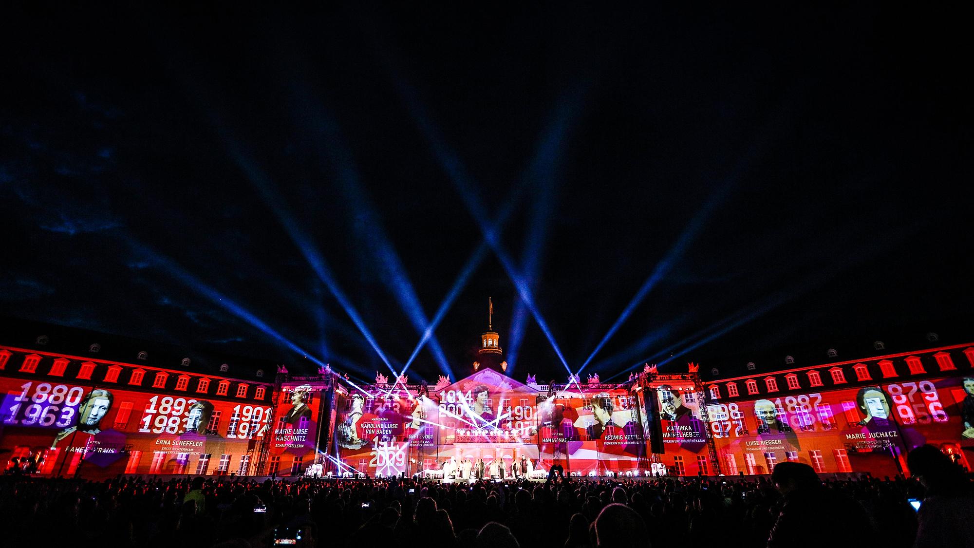Video mapping of Karlsruhe's history is projected onto a building as a large crowd watches on.