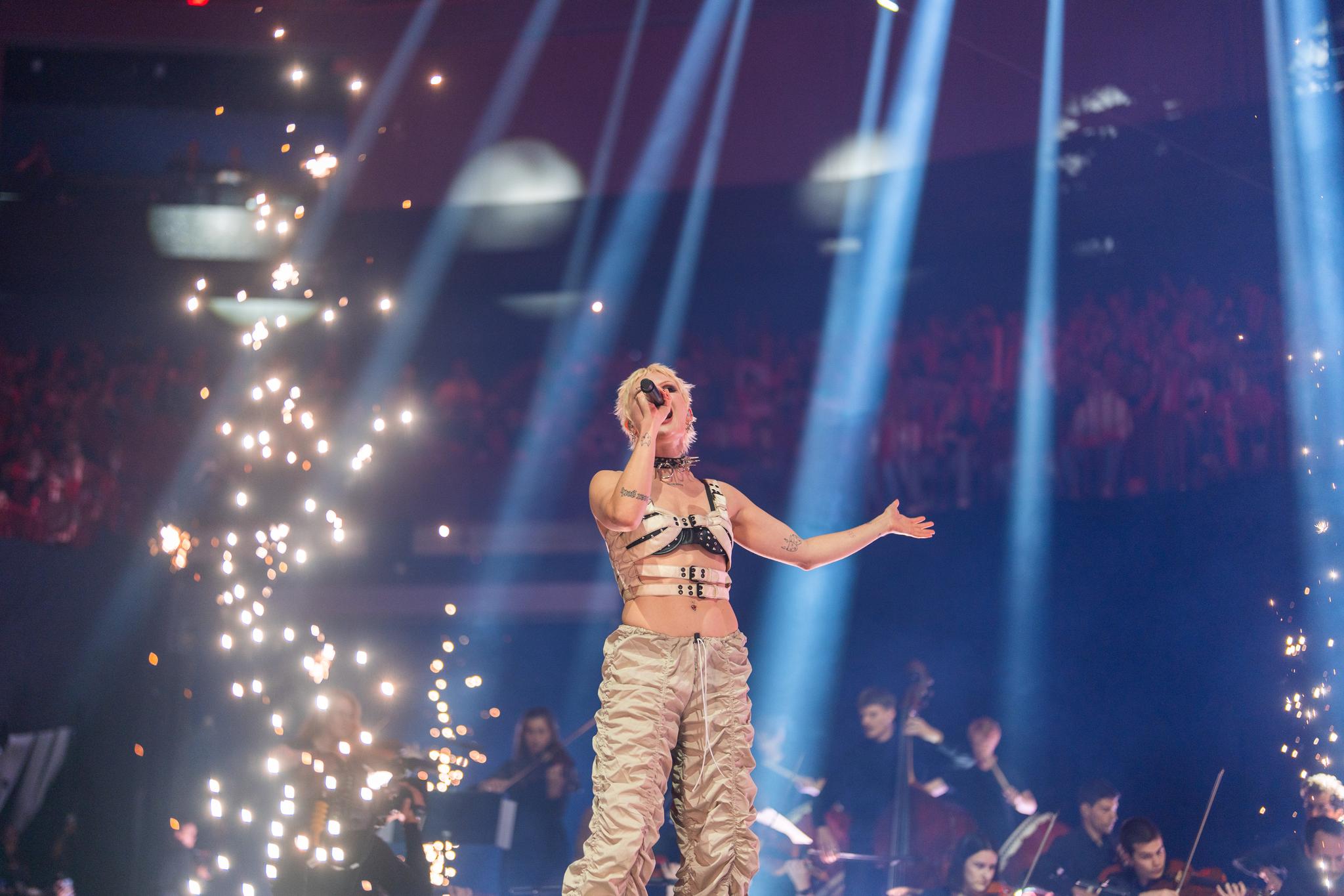 The singer Cassyette performs on stage accompanied by an orchestra and framed by sparks of pyrotechnics.