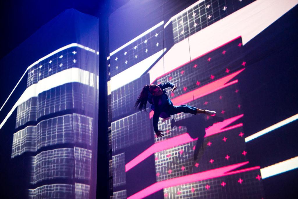 A dancer suspended from the ceiling runs across a wall projected with pink and white stripes.