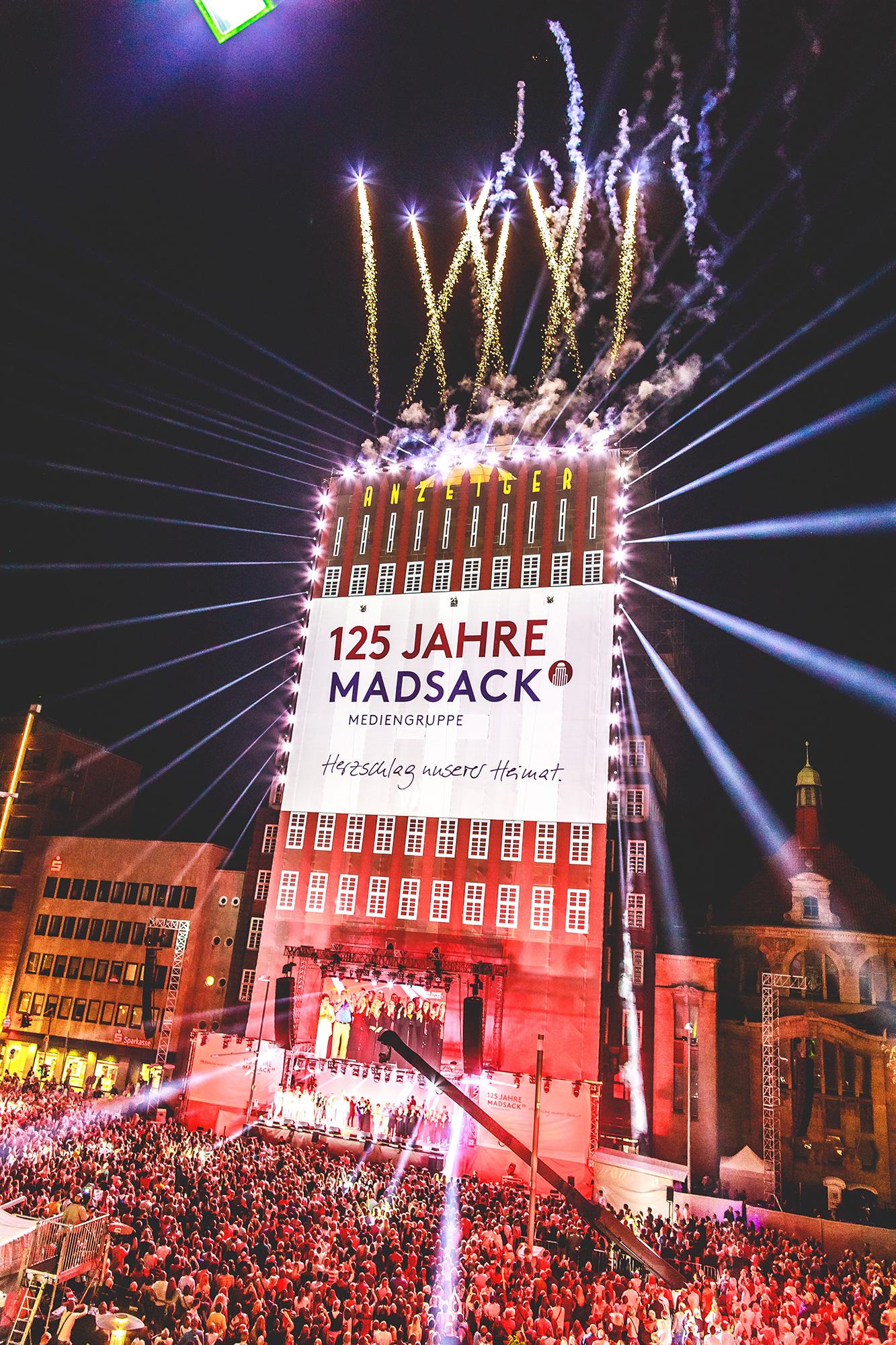 Video mapping of a building and "125 Jahre Madsack" on a large vertical stage. Fireworks burst out from the top. A large crowd watches outdoors.