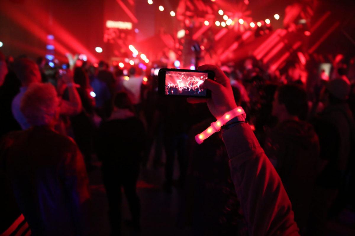 A large crowd faces a stage with performers. The scene is lit in red. A hand with a glowing armband raises a phone, which is recording the surroundings.