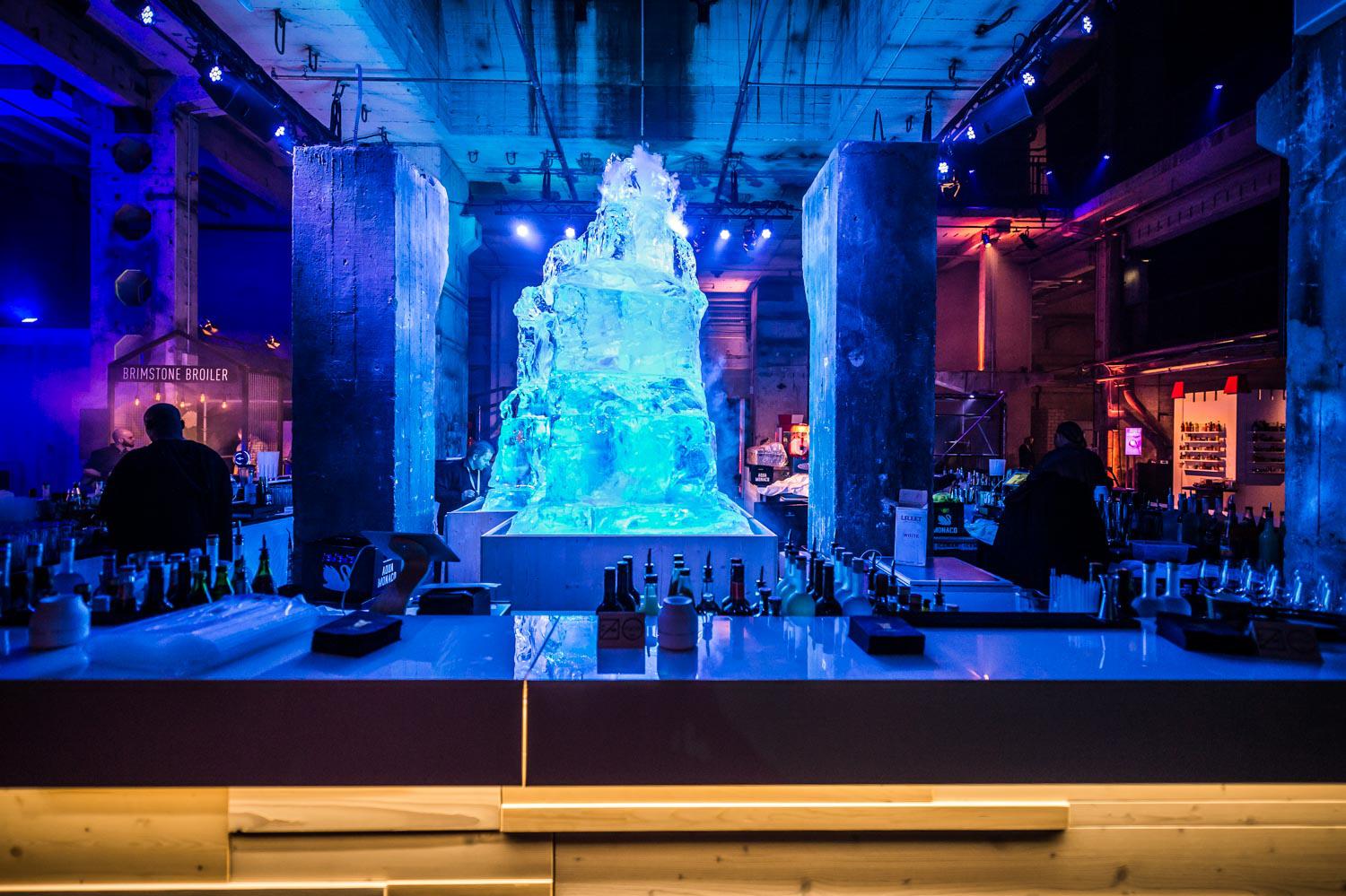 A transparent, iceberg like sculpture glows blue behind a bar in a club environment.