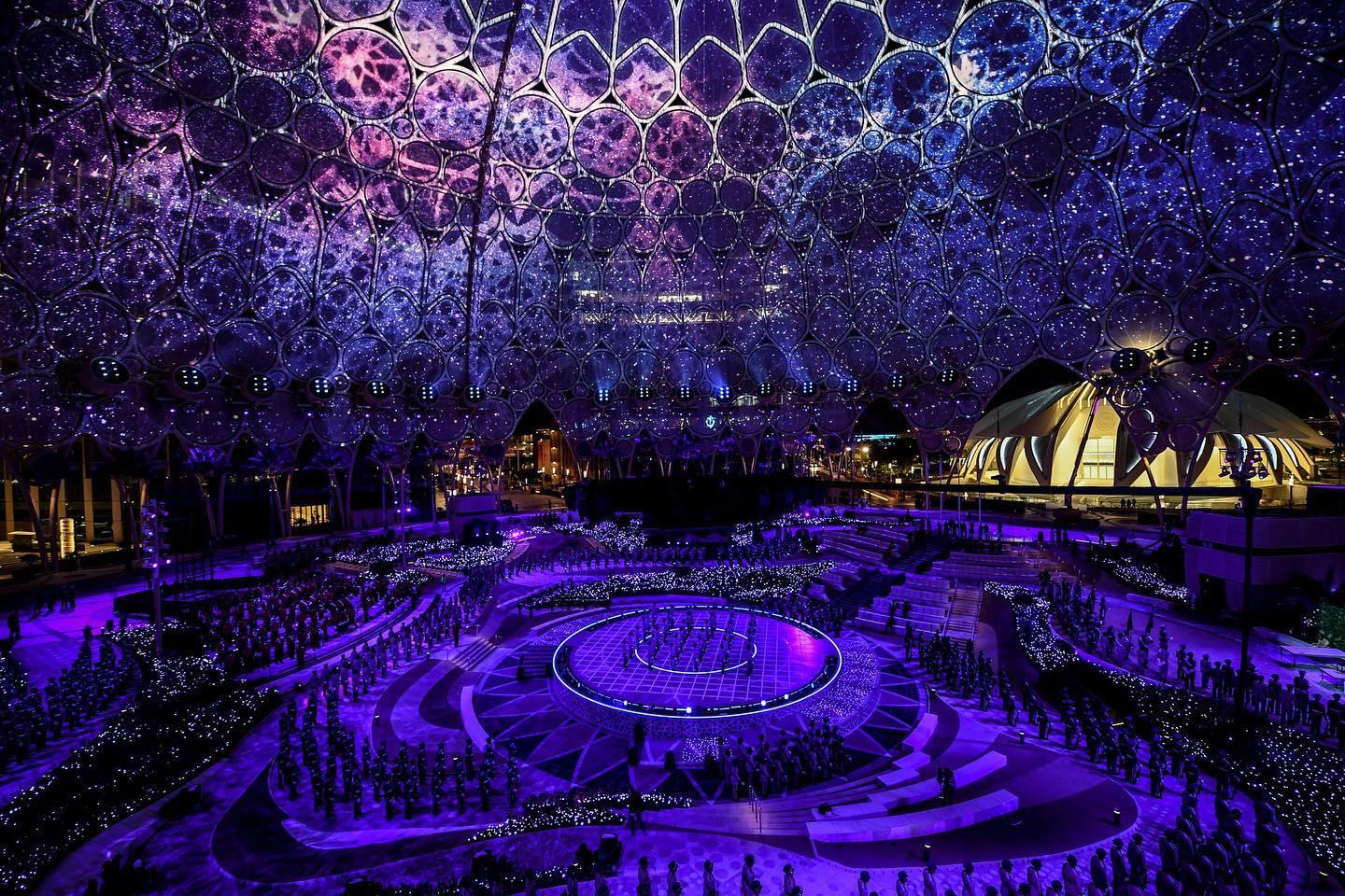 Formations of performers are under a large, glittering dome with intricate geometric patterns.