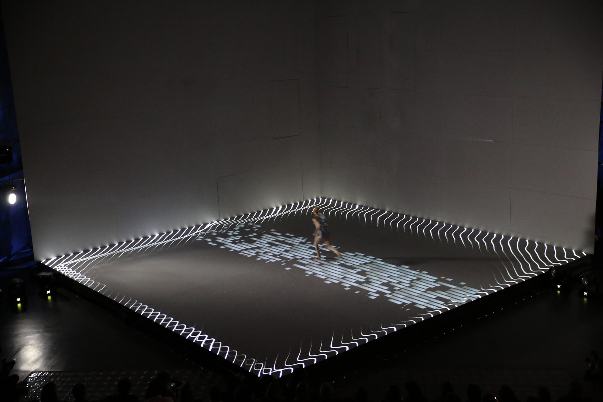 A dancer runs across the cube shaped stage with sparse projections underneath them.