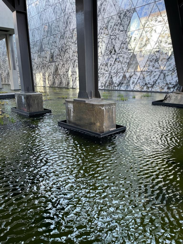 Metal beams on top of concrete blocks go into a pond. A marble wall is on the other side.