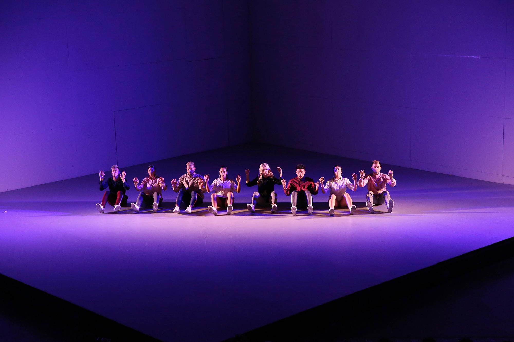 8 dancers sit in a line on the cube shaped stage with their palms out facing the audience. The stage is lit in lavender.