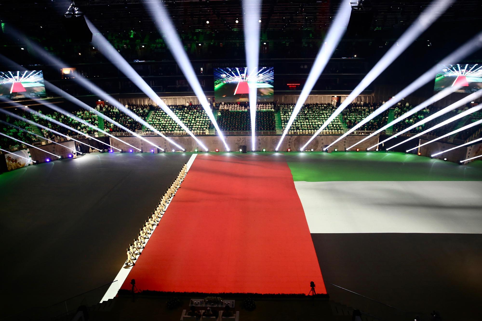 A line of police officers walk in formation across the arena. Behind them the UAE flag unfolds via projection mapping on the ground.