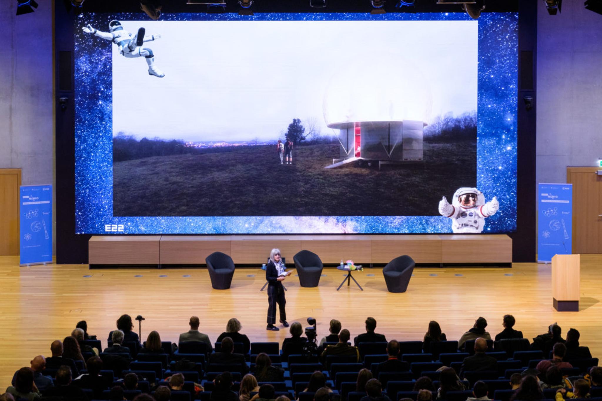 Someone gives a lecture in a modern looking lecture hall with images of astronauts behind her.