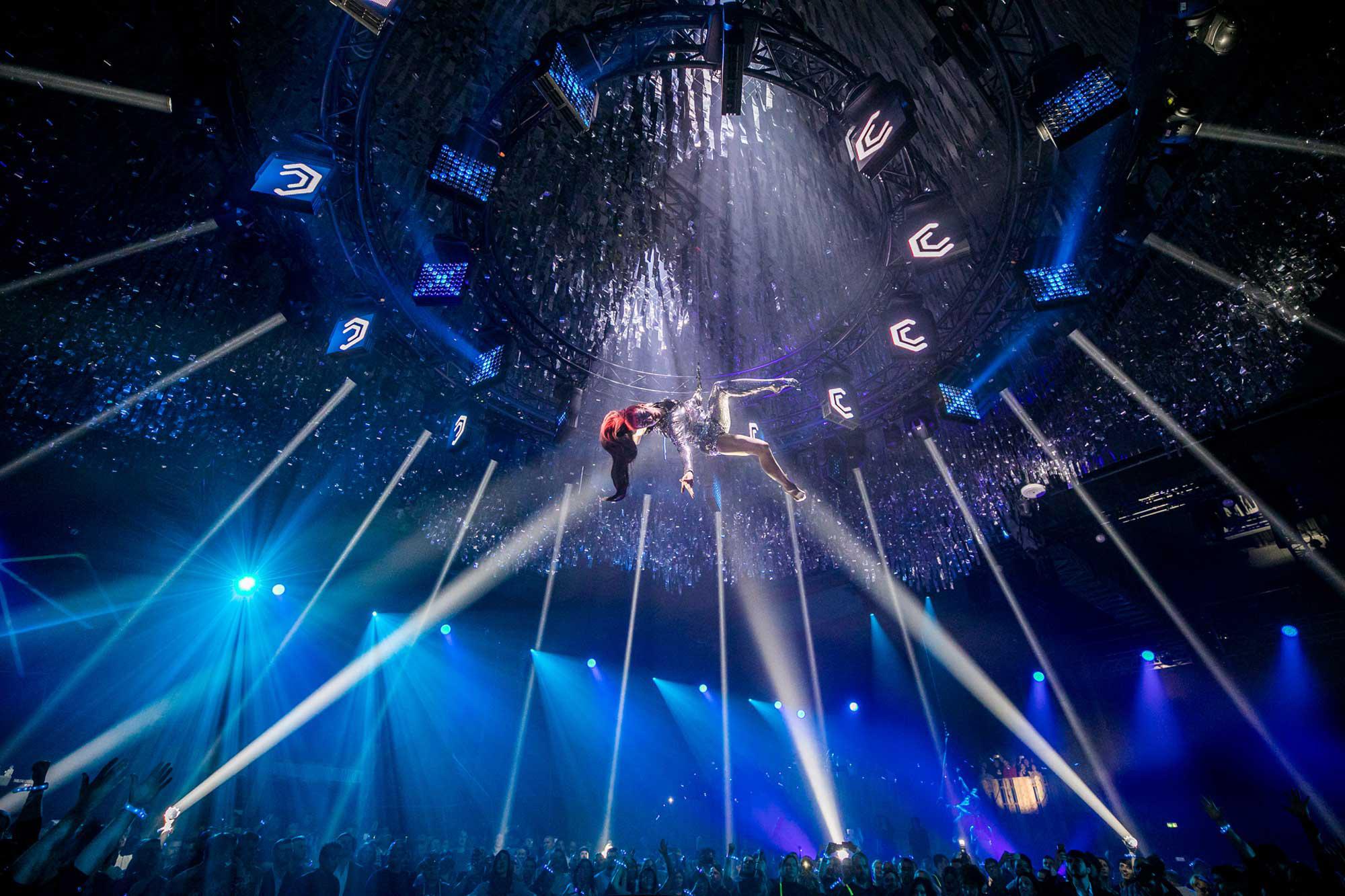 An acrobat hangs horizontally from the ceiling, descending from circular trusses with square club lights beaming from its circumference. Beams of white light shine onto the acrobat, whose long red hair and right hand reach down towards the sea of people, lit in blue, underneath her.