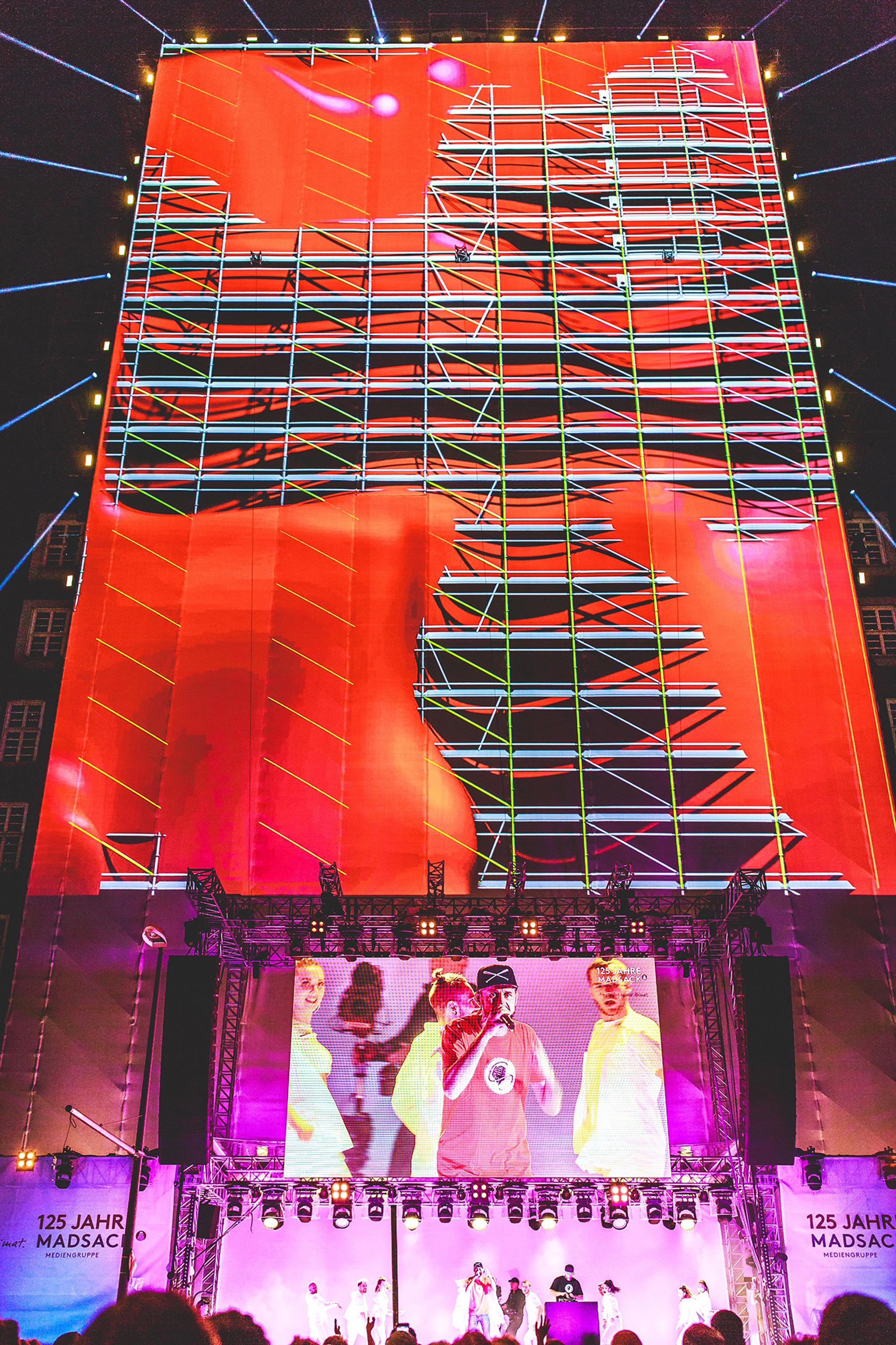 Video mapping of red splotches overlayed on a scaffolding pattern on a large vertical stage. A large crowd watches outdoors.