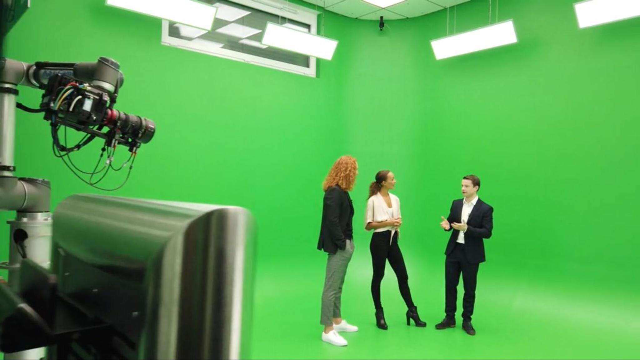 3 people stand on a greenscreen. A camera is pointing at them on a mechanical arm.