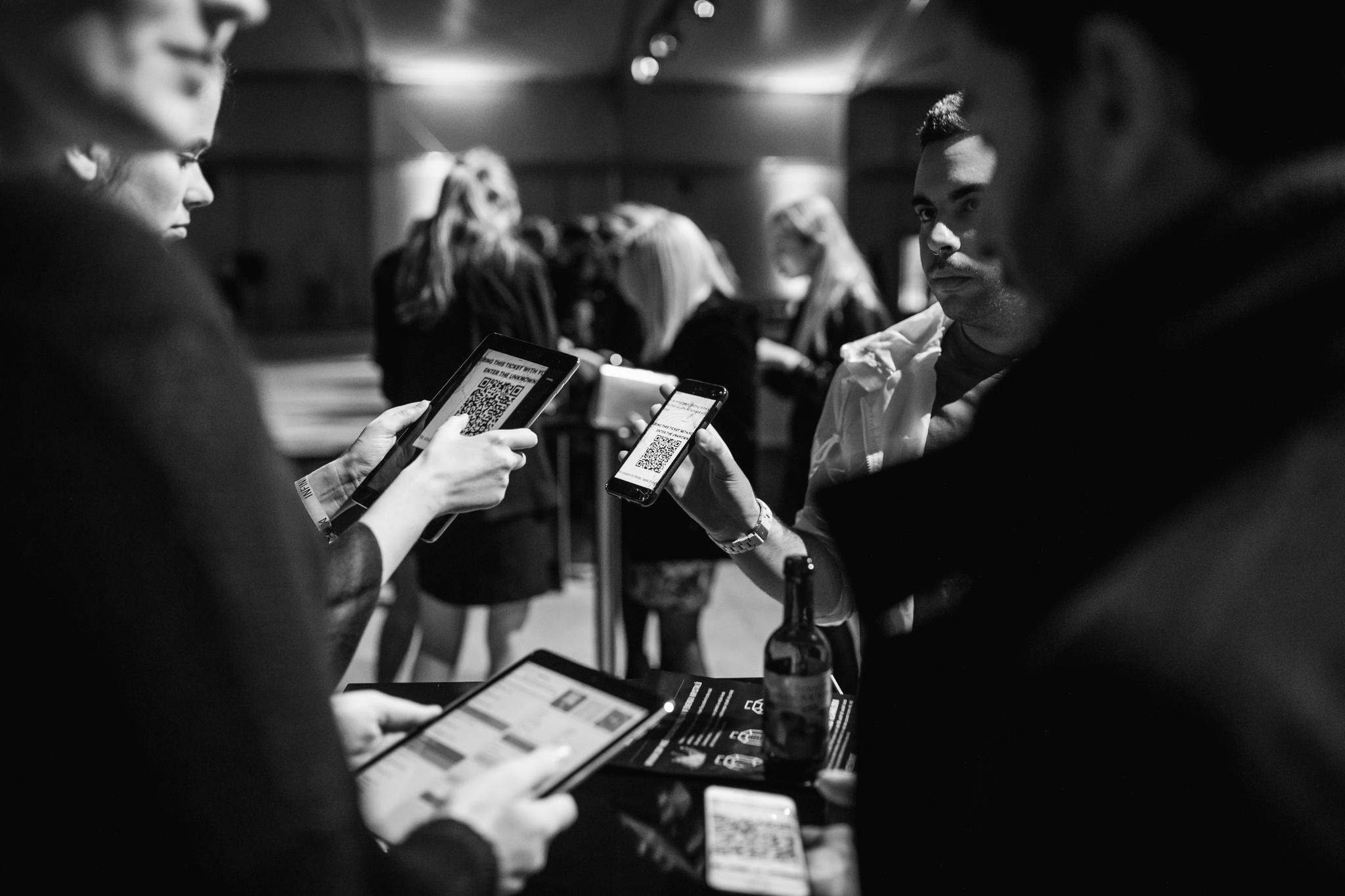 Two out of focus men frame the image left and right. The one on the left holds an ipad. A woman behind him holds another ipad with a large QR code on it. A man behind the man on the right holds out his phone to the woman; the phone also has a QR code on it. A group of women in business clothes queue in the background.