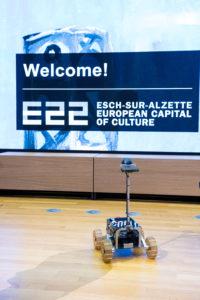 A robot on a light wooden stage. "Welcome! E22" in the background.