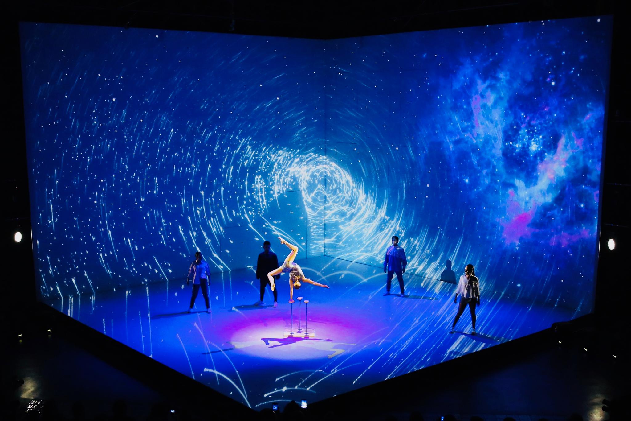 5 people are on a stage that is like a cube that's had 3 sides taken off so we can see inside of it. in the center, there is an acrobat doing a handstand with one hand on a thin, circular pedestal. The acrobat is highlighted by a spotlight, and 4 people stand far apart, framing her. The entire stage is bathed in a projection of a blue, swirling scene of outer space.