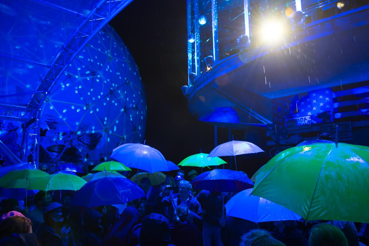 A concert takes place in blue lighting at the dome on Teufelsberg. We see the dome and a crowd of people holding decorative, purposeful umbrellas.