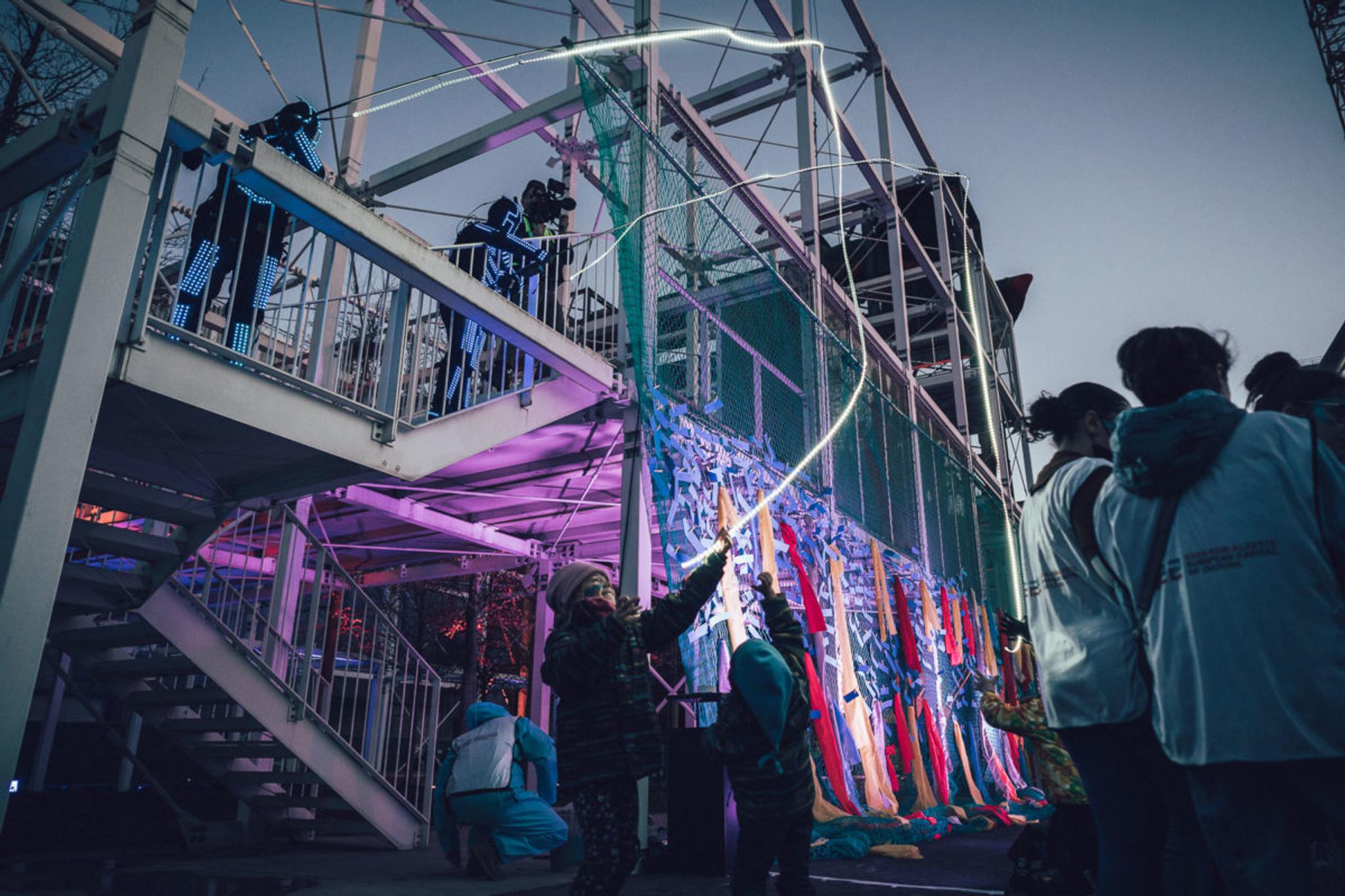 On metal scaffolding, two performers in LED suits cast two white, glowing LED fishing rods down to 2 children who excitedly play with them. A group in white hi-vis vests is in the corner- it's dusk.