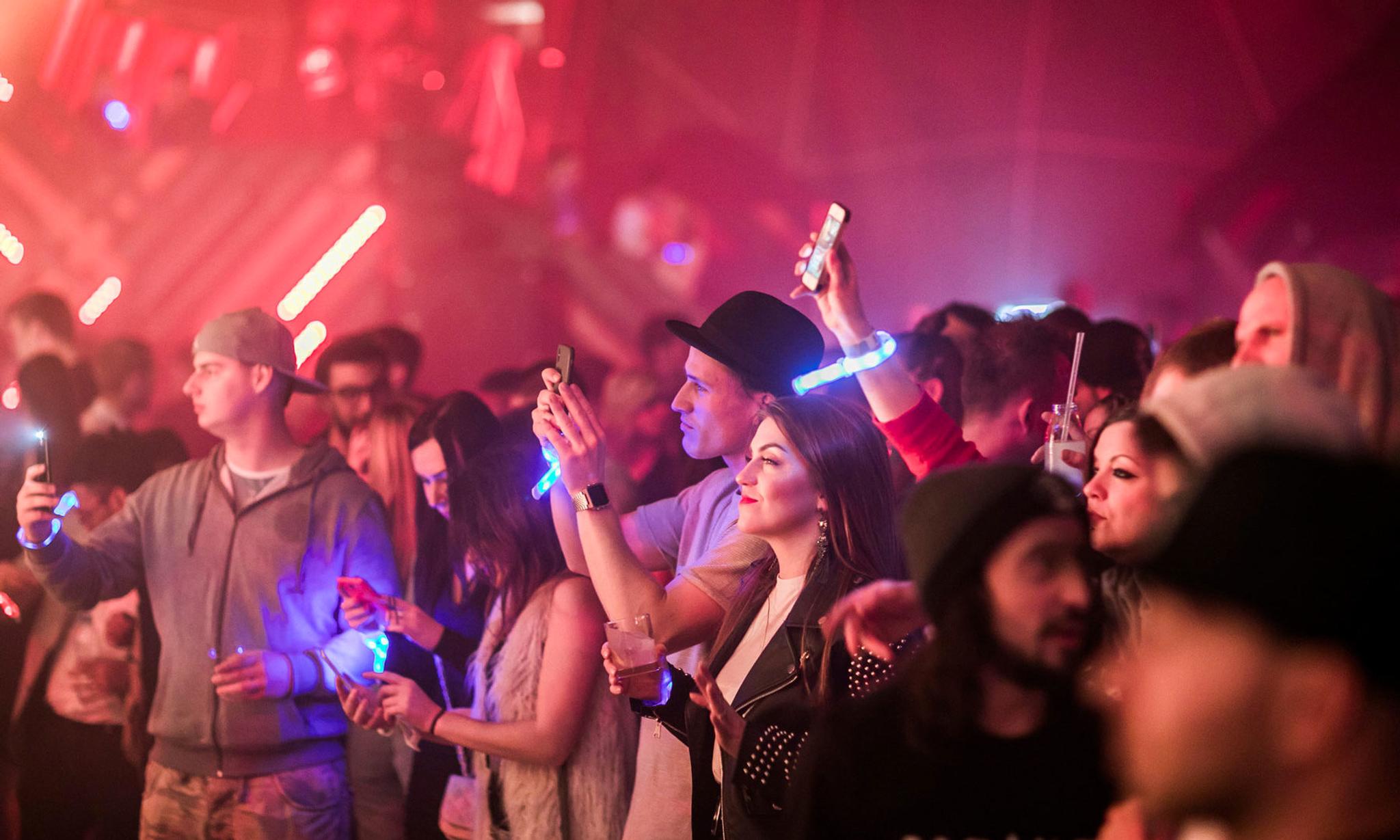 Partygoers in glowing white wristbands socialize and photograph their surroundings in a club environment.
