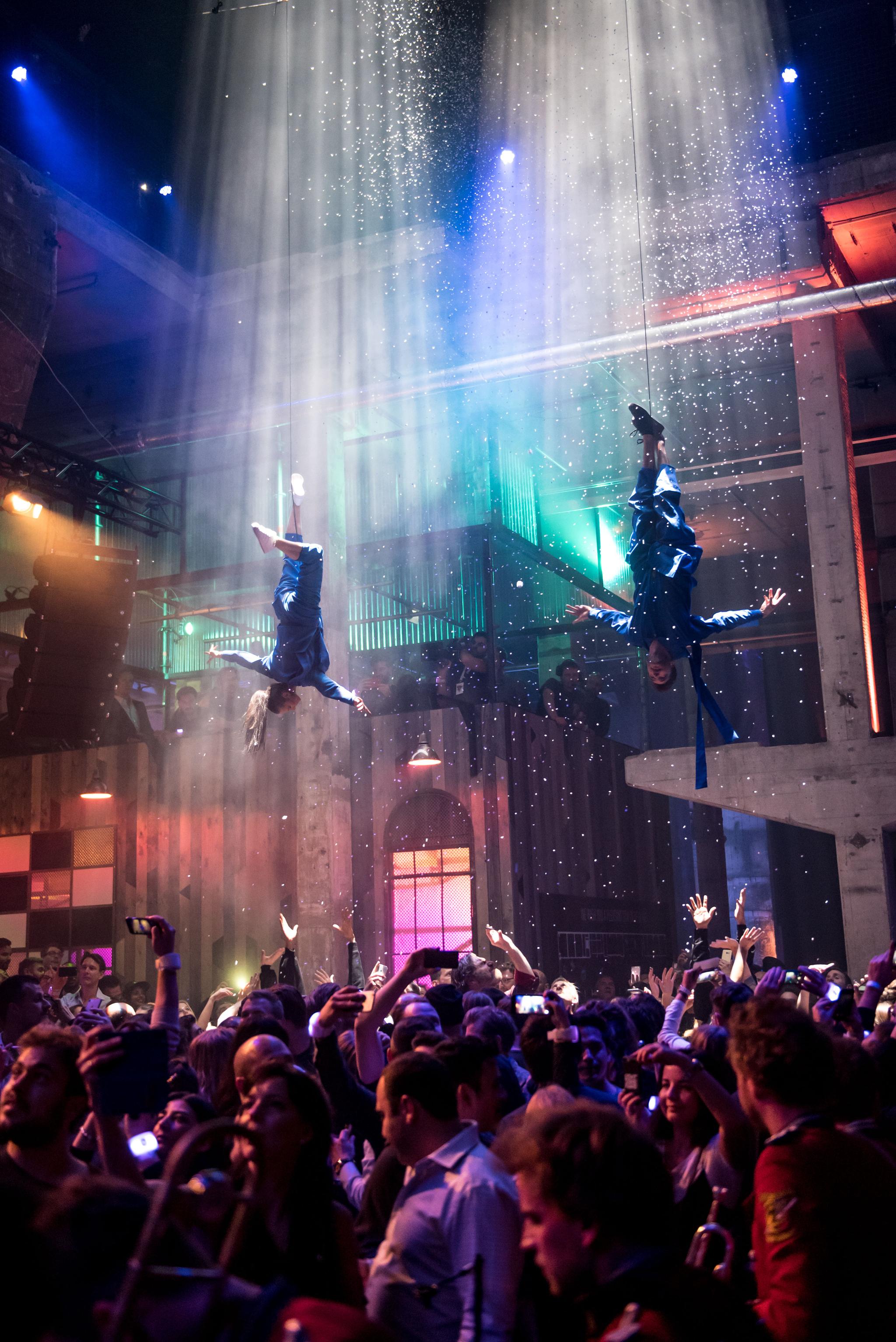 Two acrobats hang upside down from two cables, respectively, bathed in a shower of light and confetti coming from the ceiling. Below them is a lively crowd. We are in a spacious club environment with exposed concrete and high ceilings.