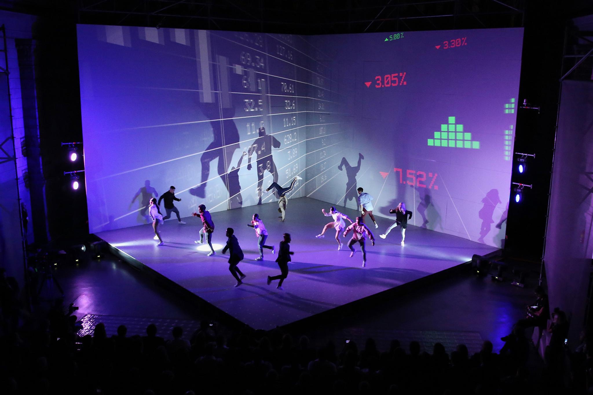 Lively dancers jump and run around the cube shaped stage where stocks are projected on the walls.