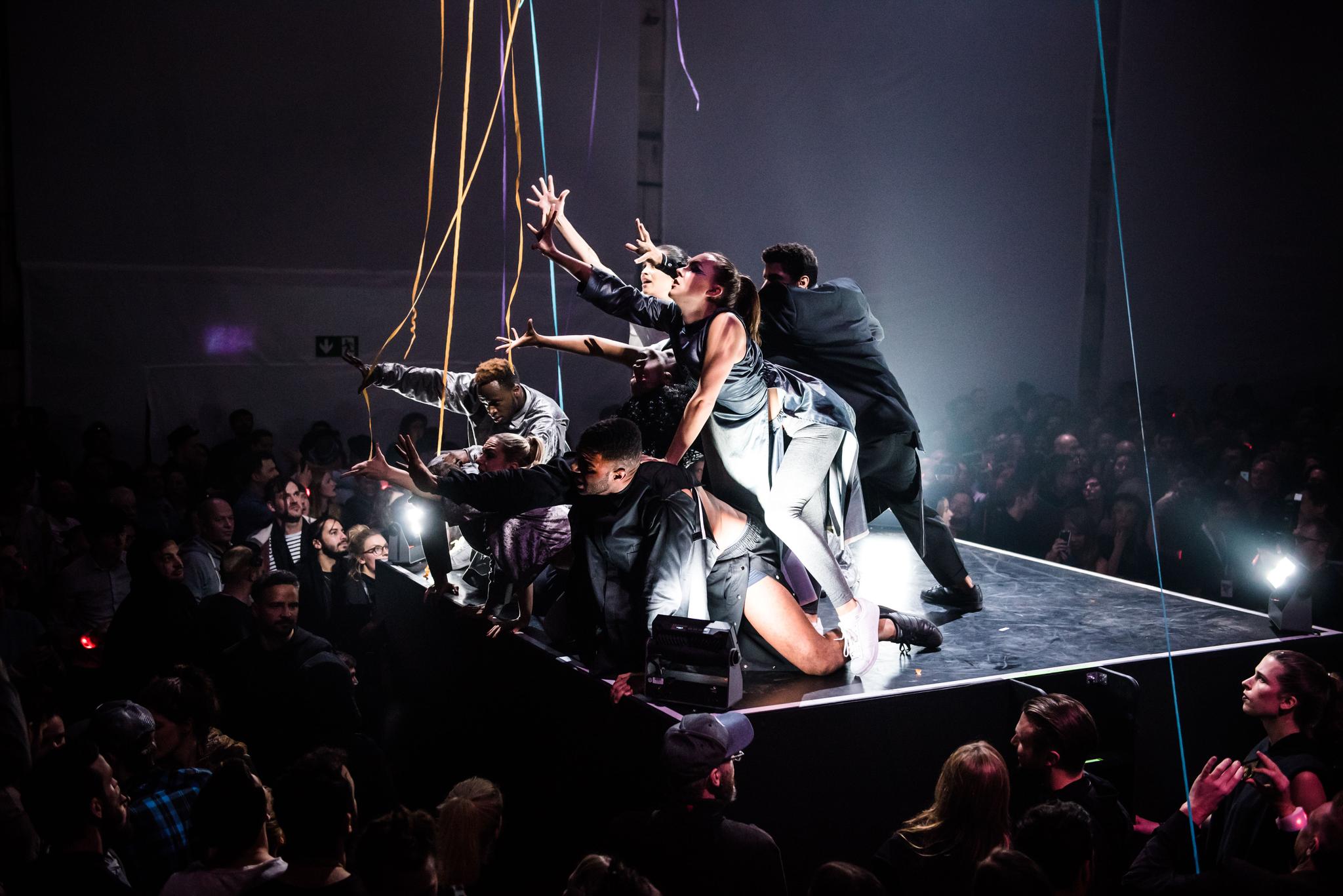 A group of dancers reach forward, out into the air, performing on a pedestal in a club setting.