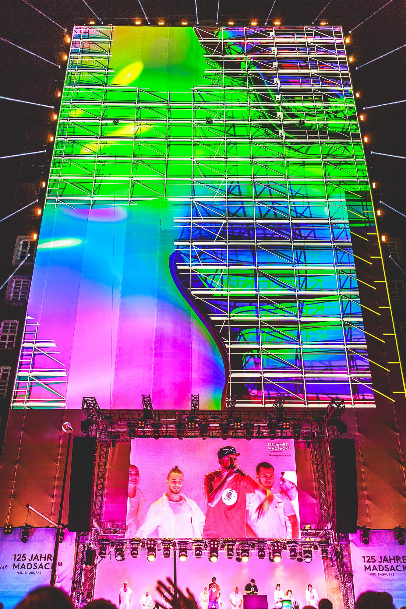 Video mapping of lava lamp like neon shapes over a scaffolding pattern on a large vertical stage. A large crowd watches a musician outdoors.