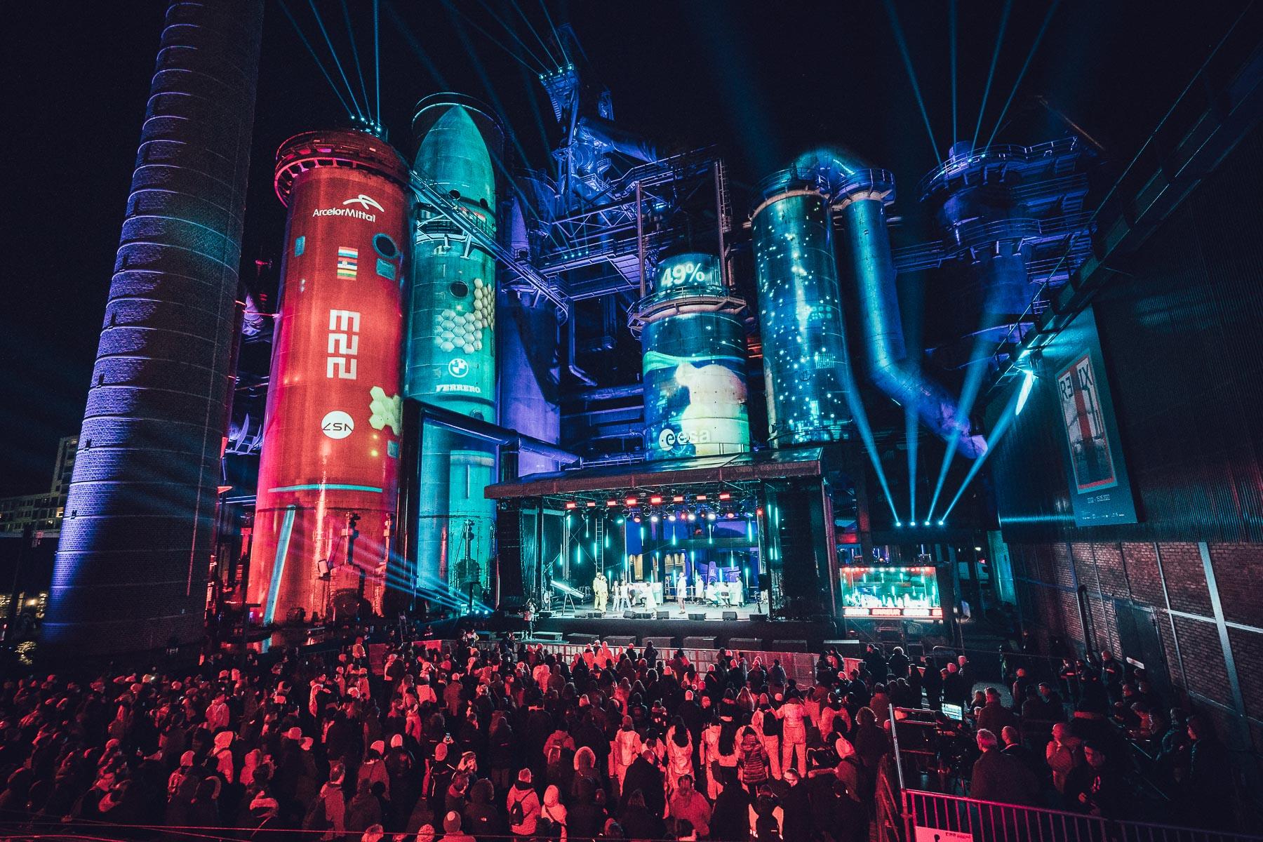 A wide shot of a crowd facing an outdoor stage. Blue lights are beaming out into the night sky, and industrial brick tube-towers and machinery frames the stage and has video content projected onto them.