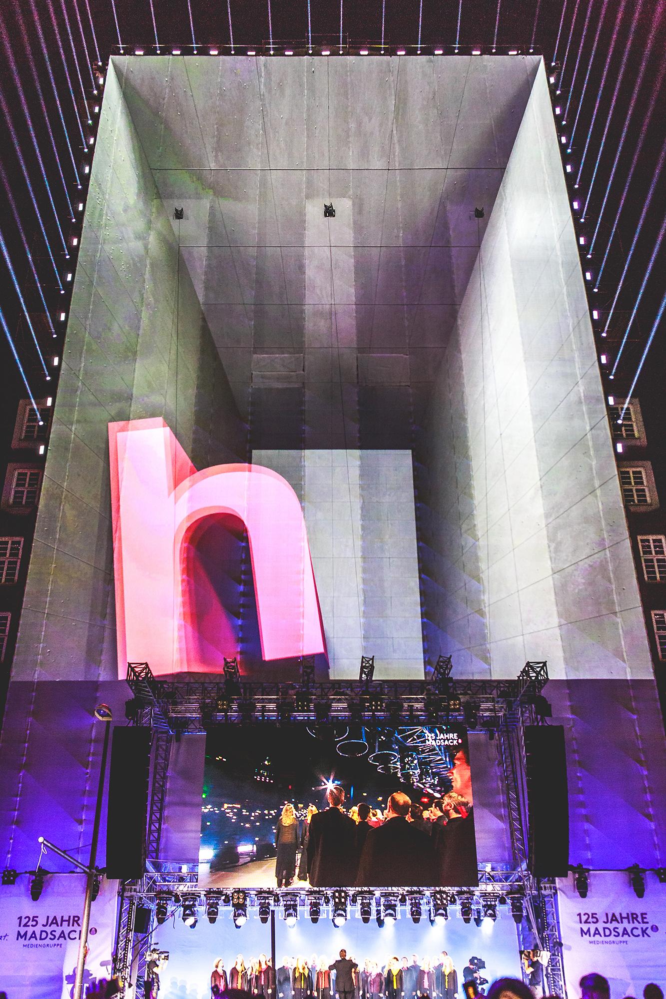 Video mapping of the letter "h" in a concrete hall on a large vertical stage. A large crowd watches outdoors.