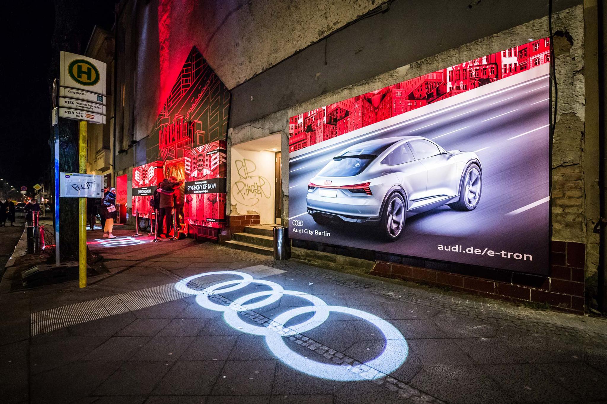The Audi logo is projected onto the sidewalk in front of an Audi ad in front of the Symphony of Now screening.