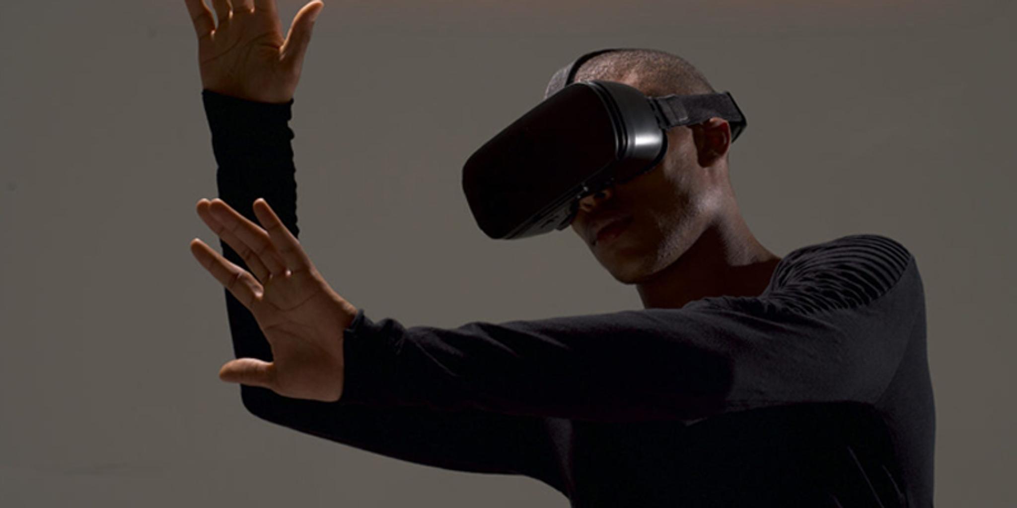A man reaches out, mime-like in front of him. He is wearing VR glasses and is photographed on a grey background.