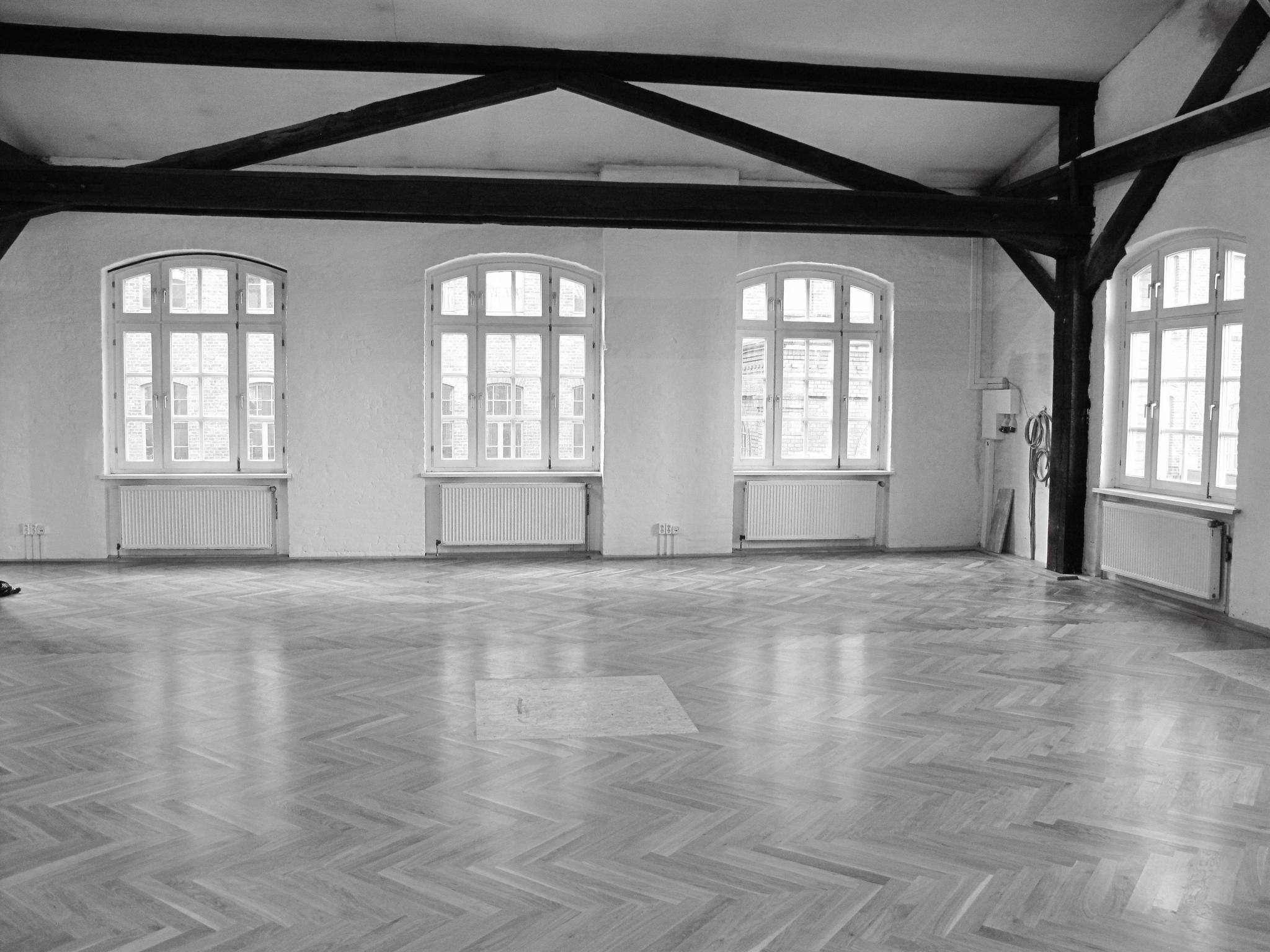 A wide shot of the studio space. Ambient light comes in through 4 arched altbau windows and dark wooden beams frame the high, triangular ceiling. The light from the windows reflects off of clean zigzagged parquet flooring.