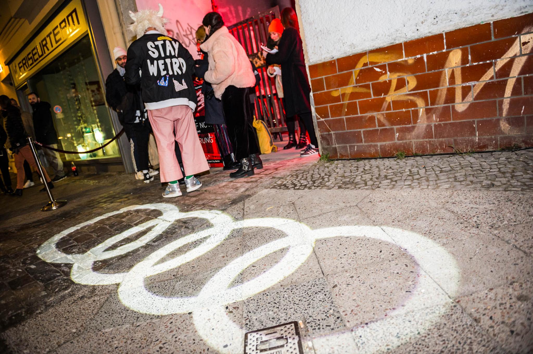 The Audi logo, 4 horizontal overlapping white circles, are projected onto the sidewalk. A yellow tag on the wall reads, hastily written, "FBM." A group of about 5 people stand outside of the door to the event. There is a black velvet rope designating where the queue is. A person in pink trousers, a blazer with "stay weird" written on the back, and a fuzzy hat with horns on it, sticks out from the crowd. We are next door to a salon.
