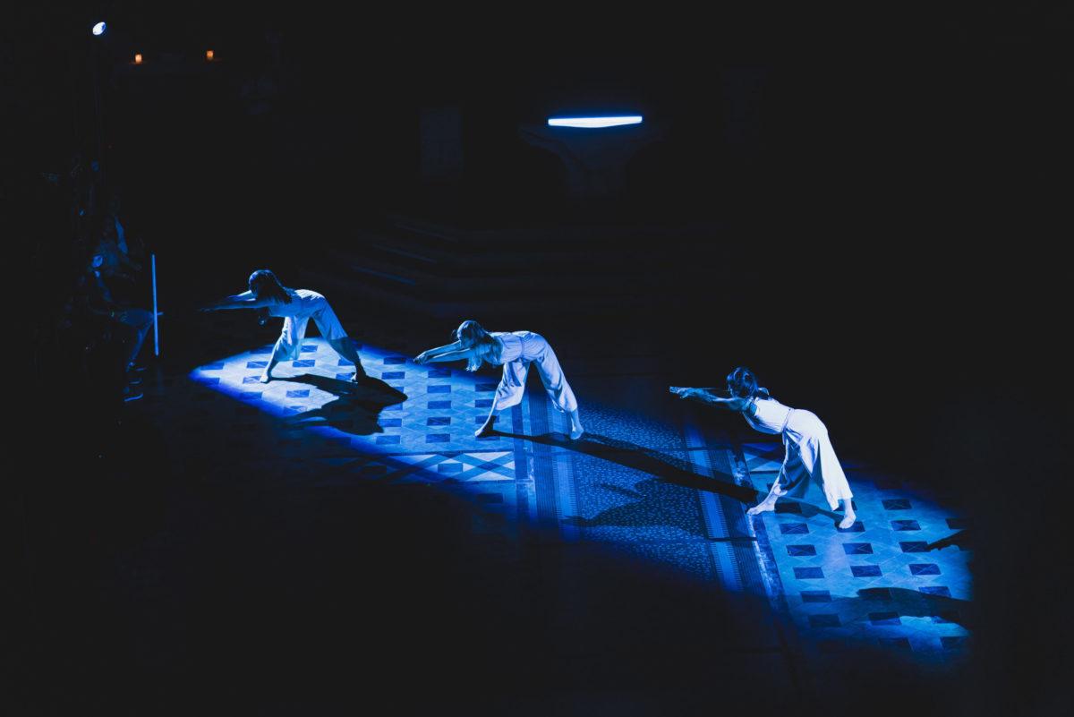 3 dancers perform in a blue shaft of light in a cathedral.