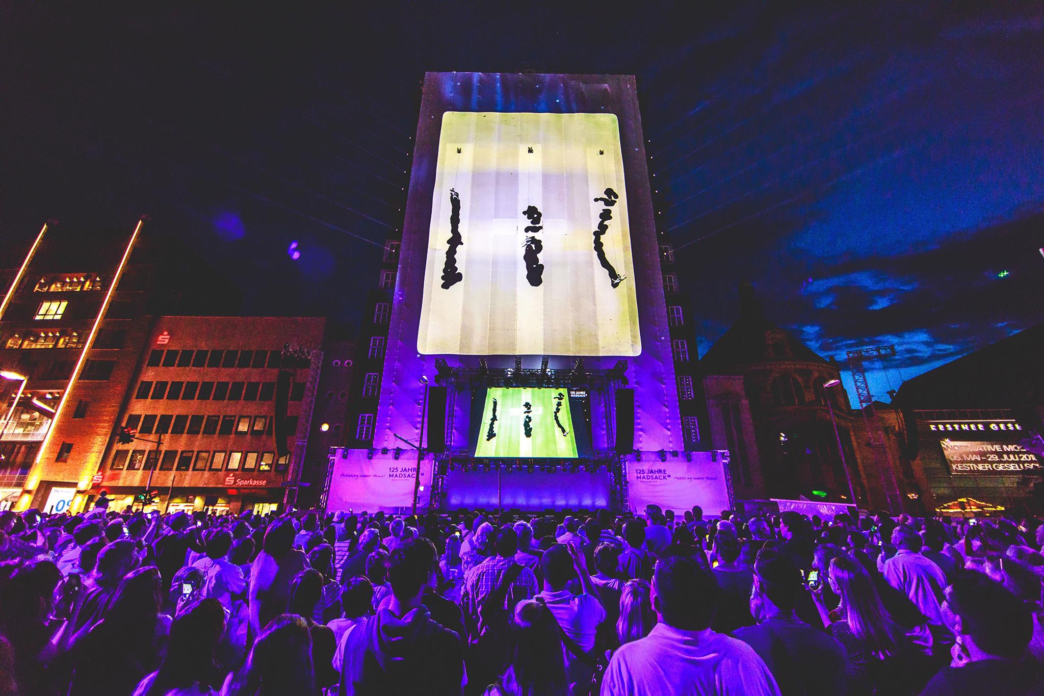 Video mapping of 3 expressionist black lines on a large vertical stage. A large crowd watches outdoors.