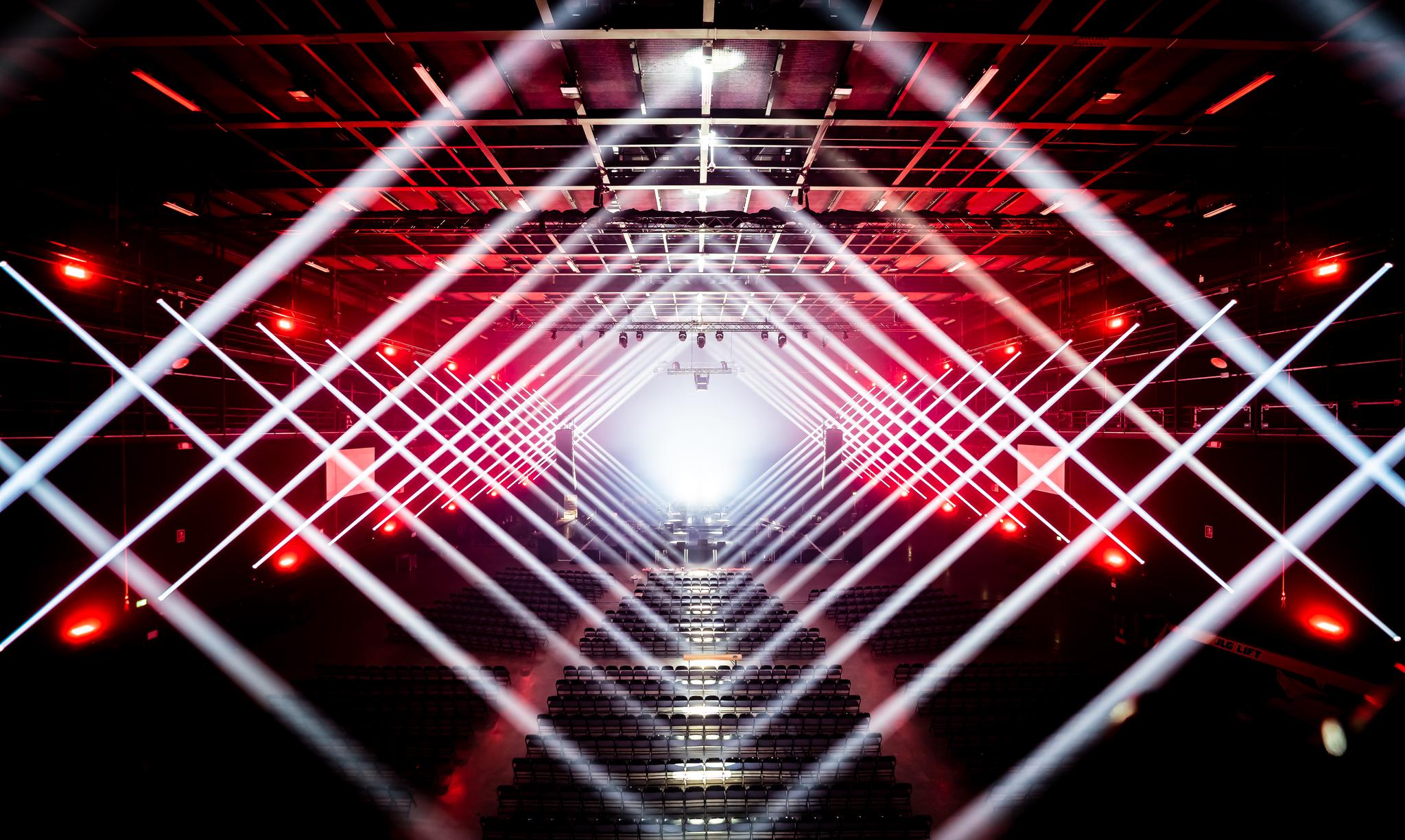 Beams of white light criss cross across an empty red-lit concert hall, forming a multi layered, uniform diamond shape.