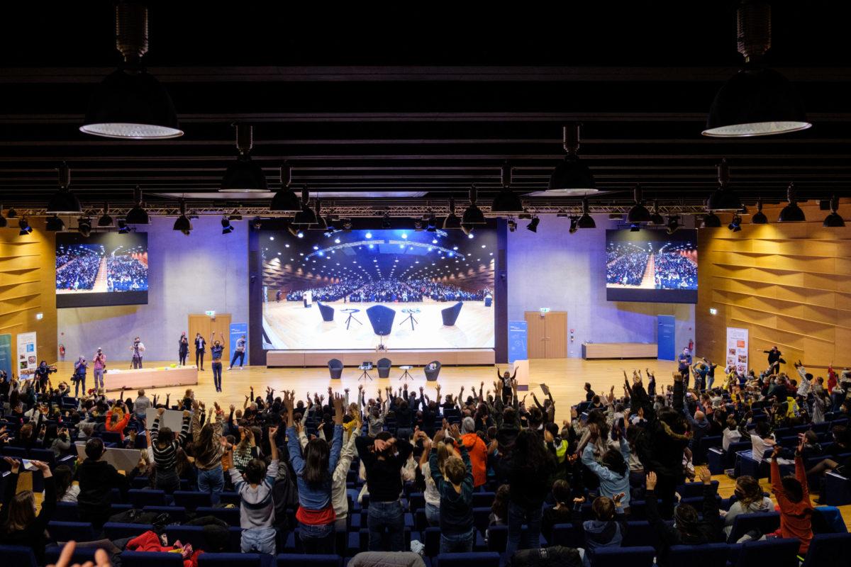 Participants in a lecture hall stand up with their hands in the air.