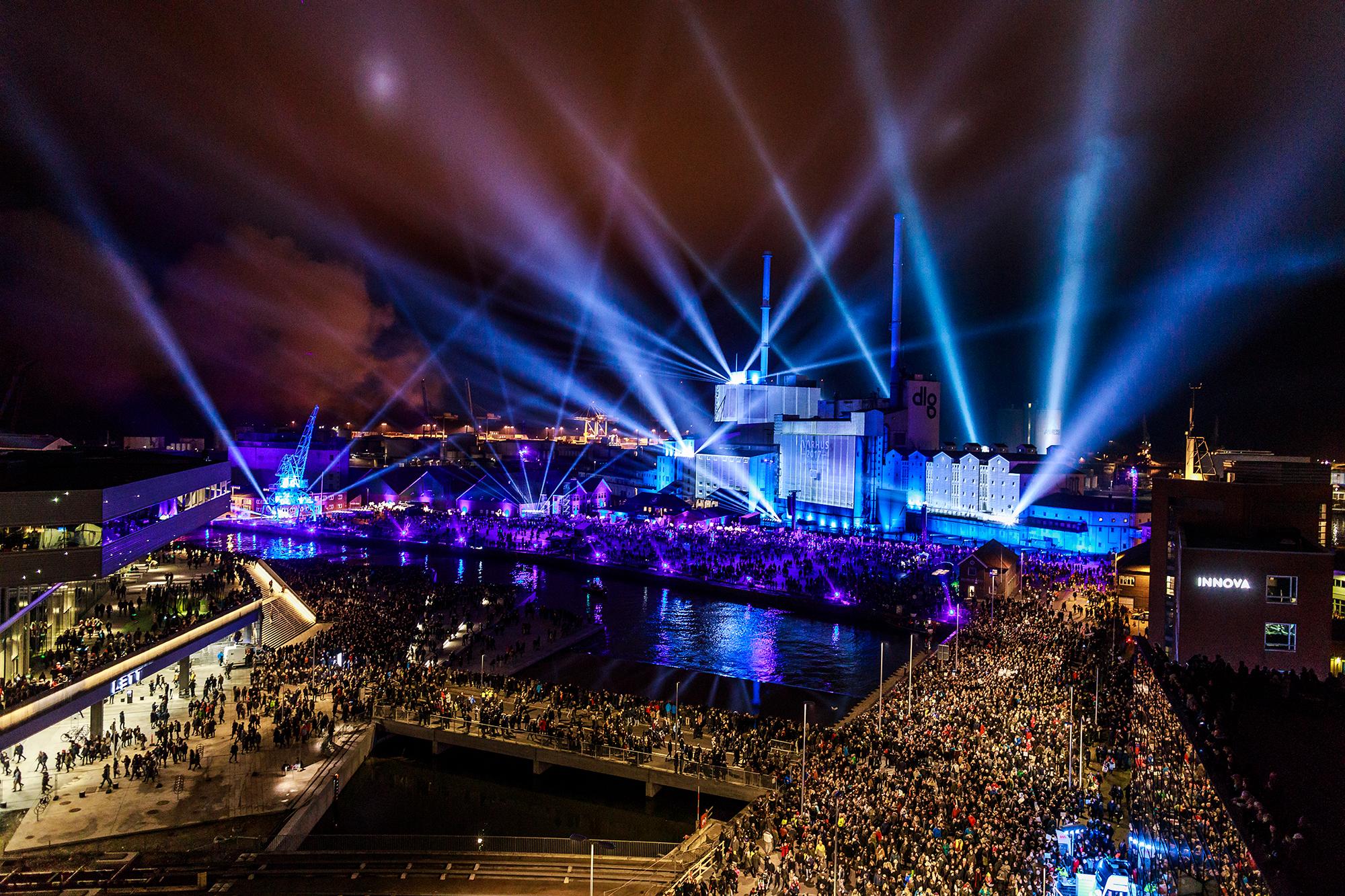 A large crowd fills the streets leading up to a building lit up in blue with spotlights shining from it.