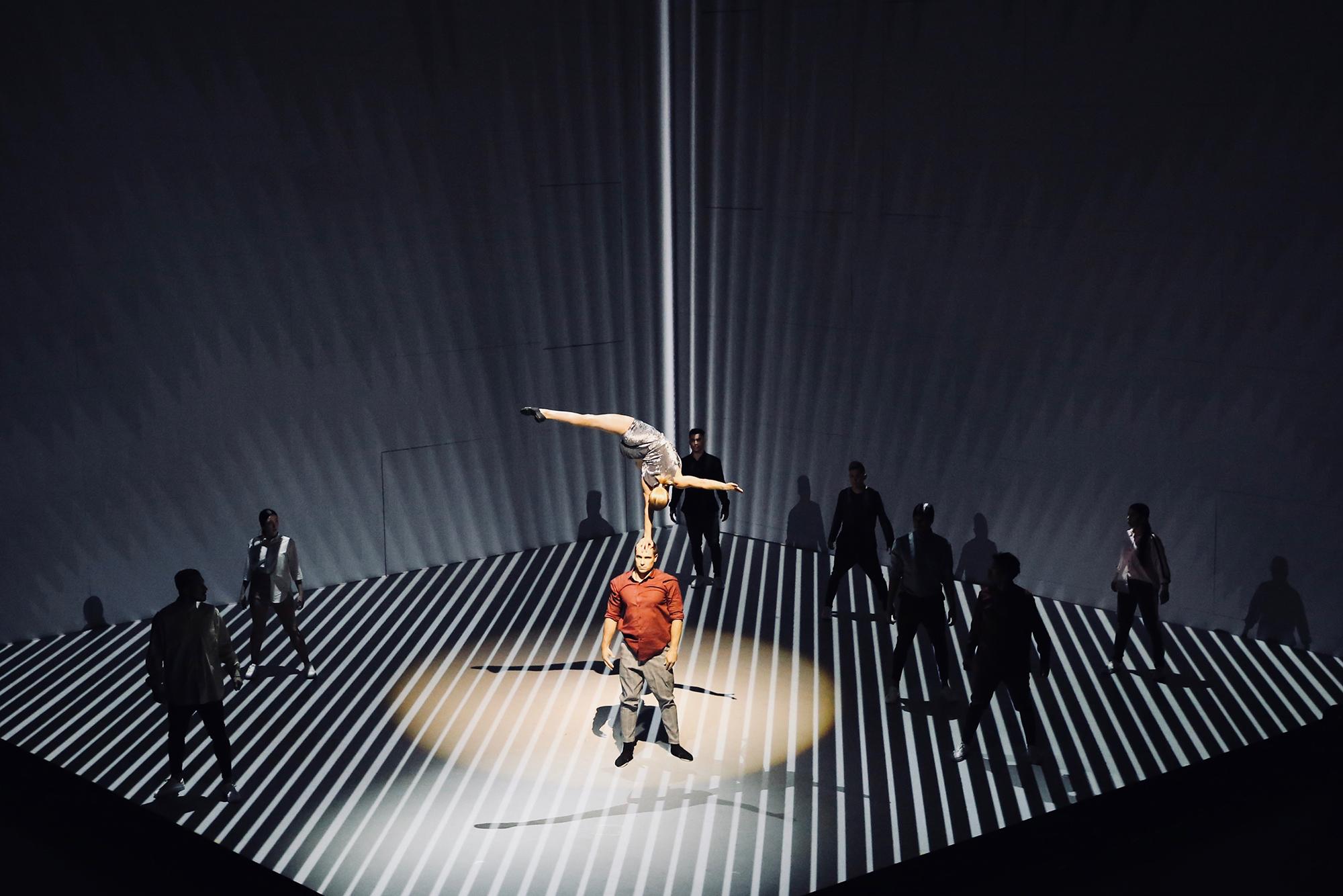 An acrobat does a one handed handstand on a man's head while other dancers look on. They all are on a cube shaped stage with stripes projected on the floor.