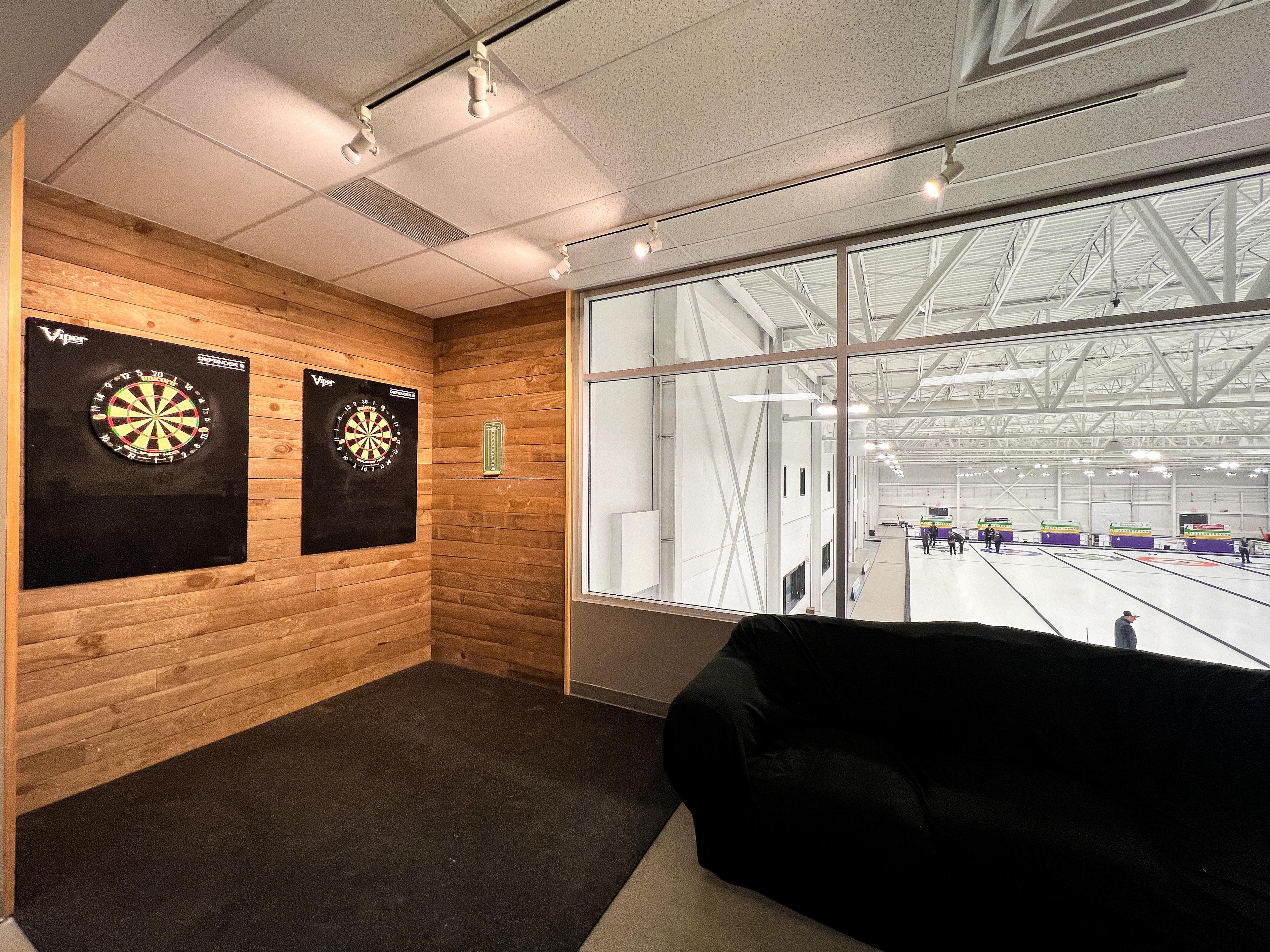 An image of a corner of the lounge with 2 dartboards hung on the wall and a couch. The back of the couch is facing a glass wall that's facing a curling rink.